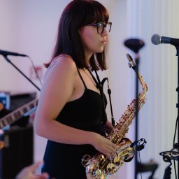 Holiday zespół muzyczny weselny obsługa wesel eventów - Saksofonistka w czarnej sukience, z okularami, grająca na saksofonie podczas występu na tle kolumny i mikrofonu. W tle widoczna gitara.