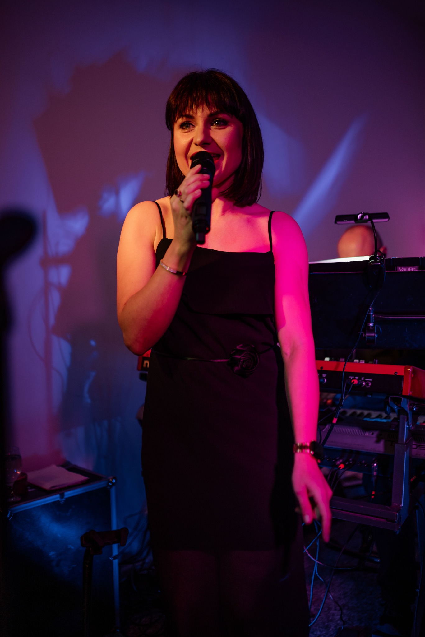 Wokalistka w czarnej sukience śpiewa do mikrofonu na scenie z instrumentami klawiszowymi w tle, oświetlona czerwonym i niebieskim światłem.