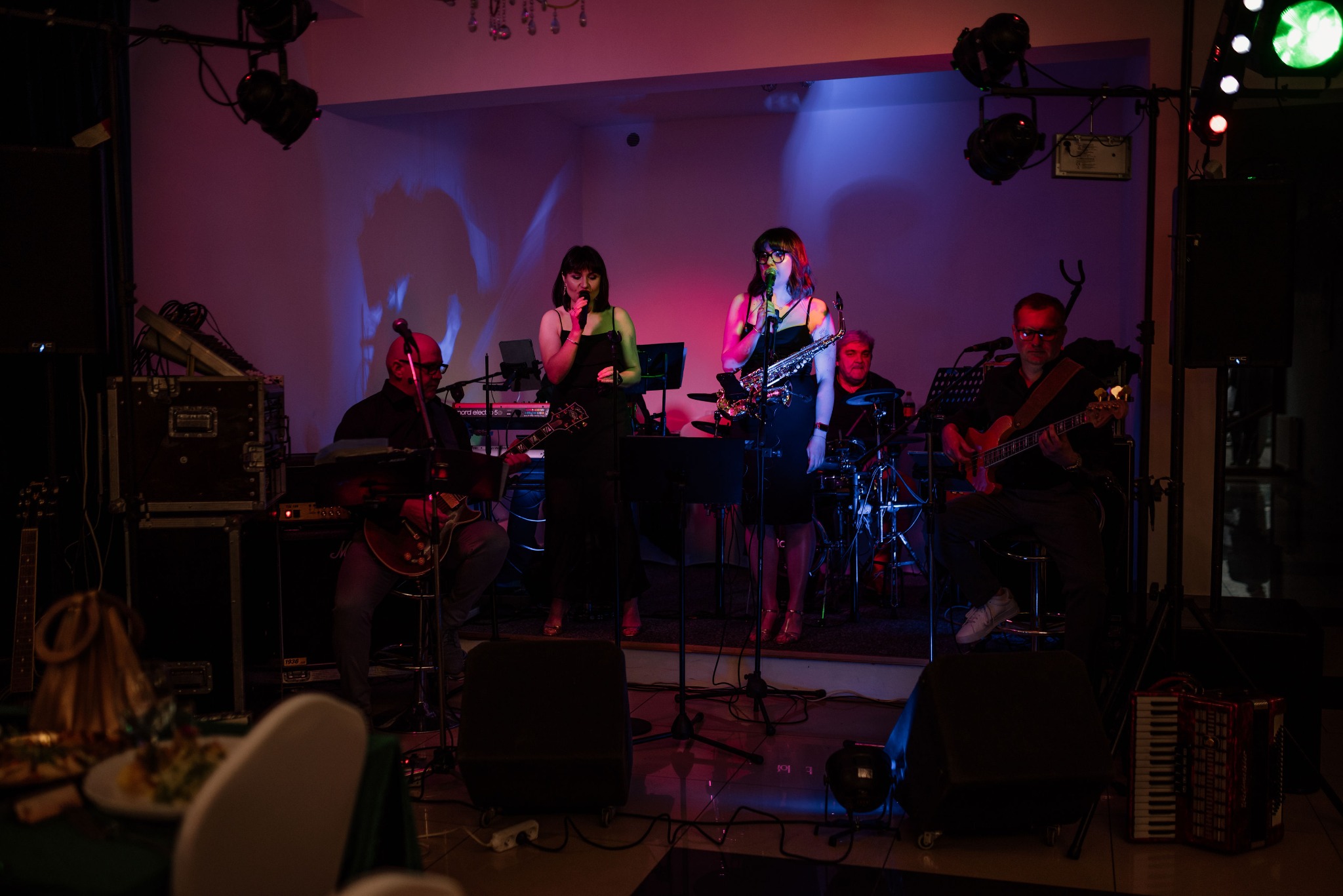 Zespół muzyczny na scenie: dwie wokalistki z saksofonem i mikrofonem, gitarzysta, basista i perkusista, oświetlenie sceniczne w kolorach różu i fioletu.