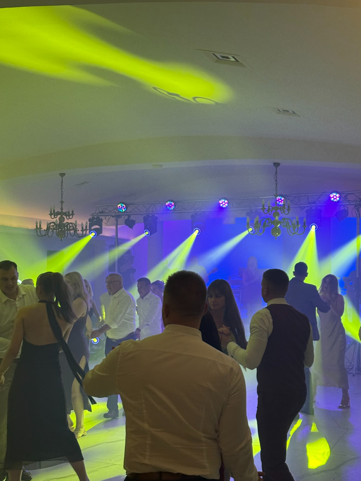 Parkiet taneczny wypełniony tańczącymi osobami, oświetlony dynamicznymi światłami scenicznymi w odcieniach żółci i fioletu, tworzącymi żywą atmosferę imprezy.