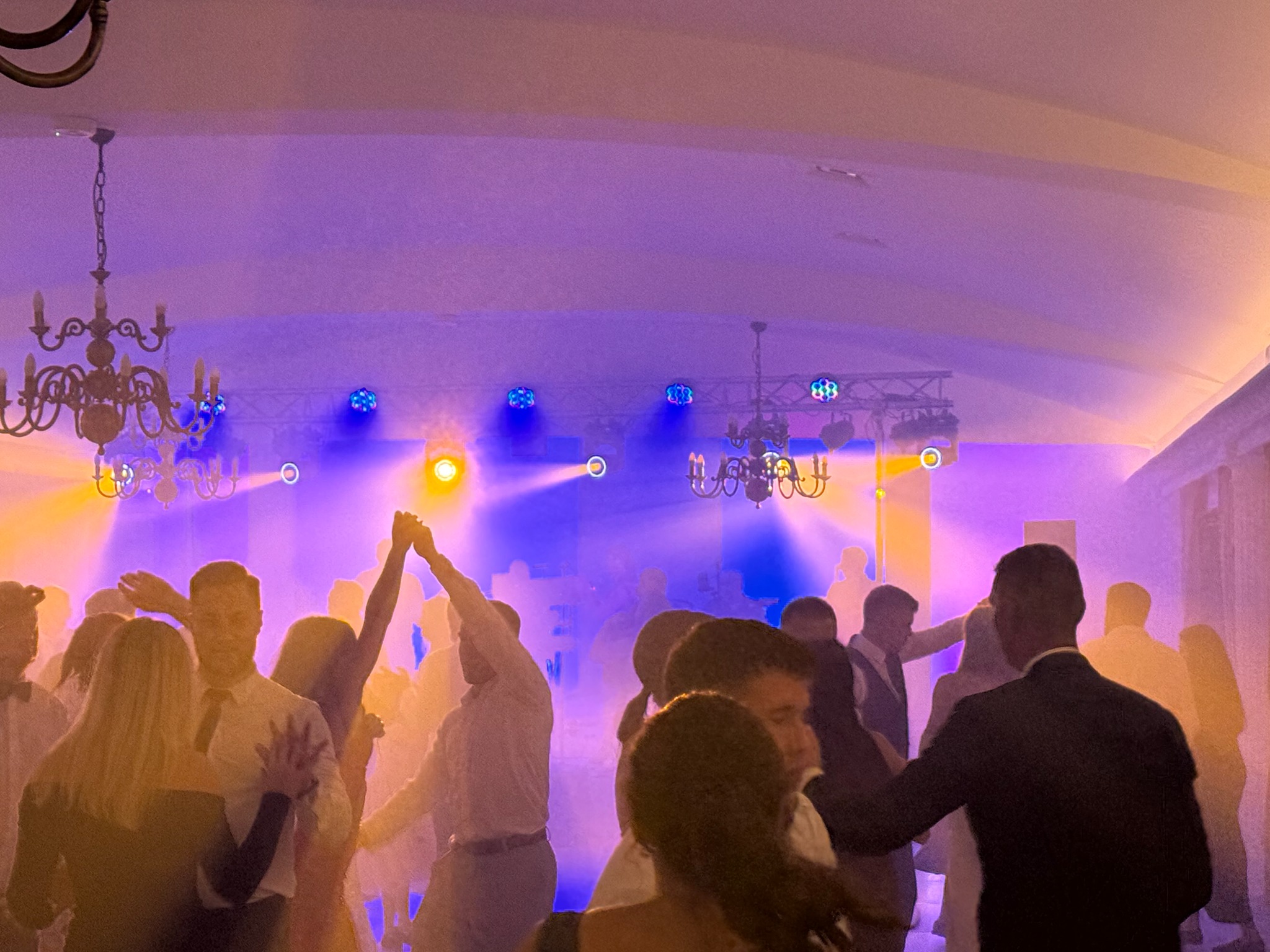 Roztańczony tłum w sali balowej, oświetlonej przez dekoracyjne żyrandole i kolorowe światła sceniczne, z efektem mgły. Uczestnicy w strojach wieczorowych.