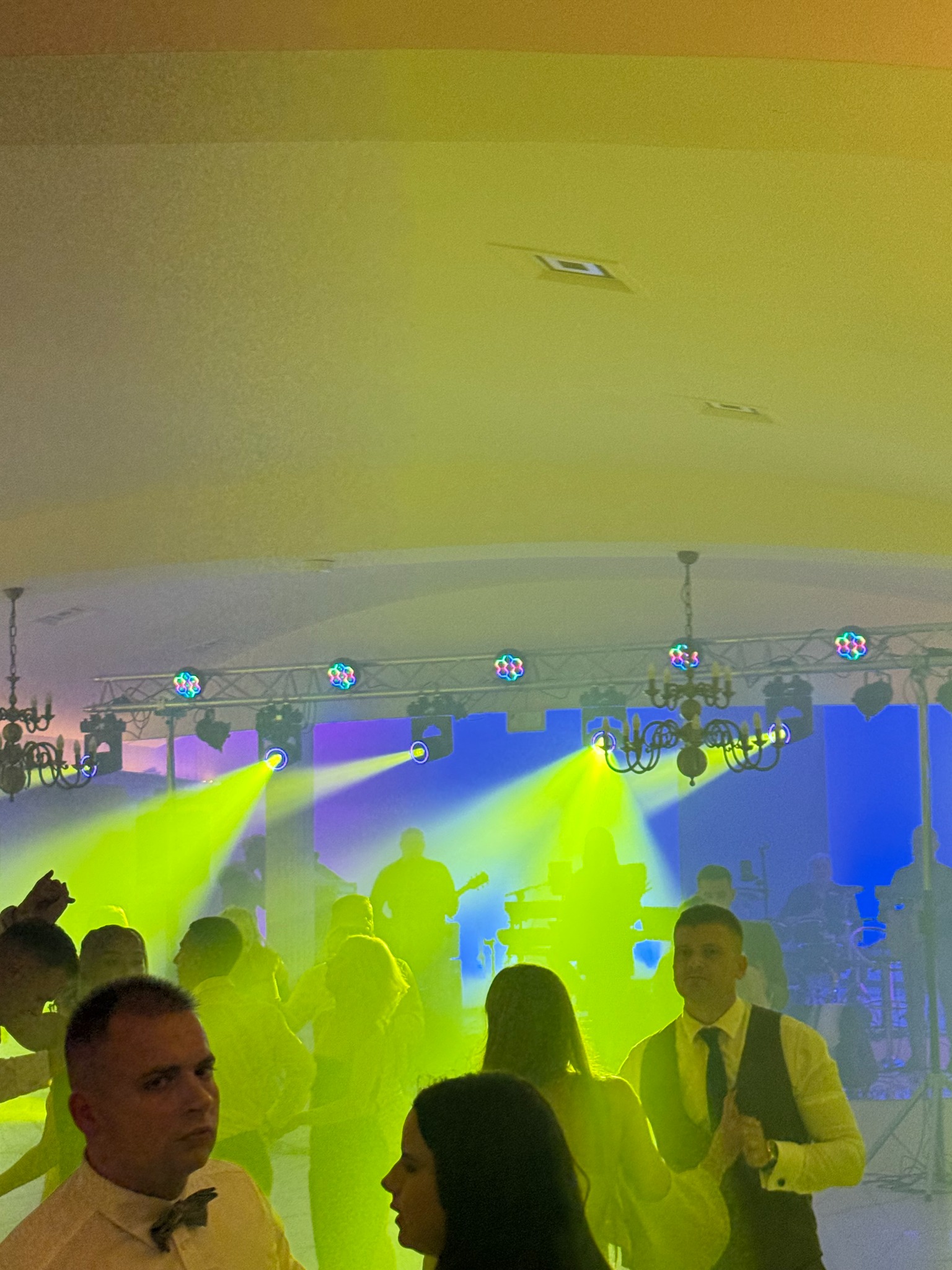 Zespół gra na scenie podczas imprezy, oświetlony reflektorami i kolorowymi światłami. W tle tańczący goście, w pierwszym planie widoczni uczestnicy zabawy.