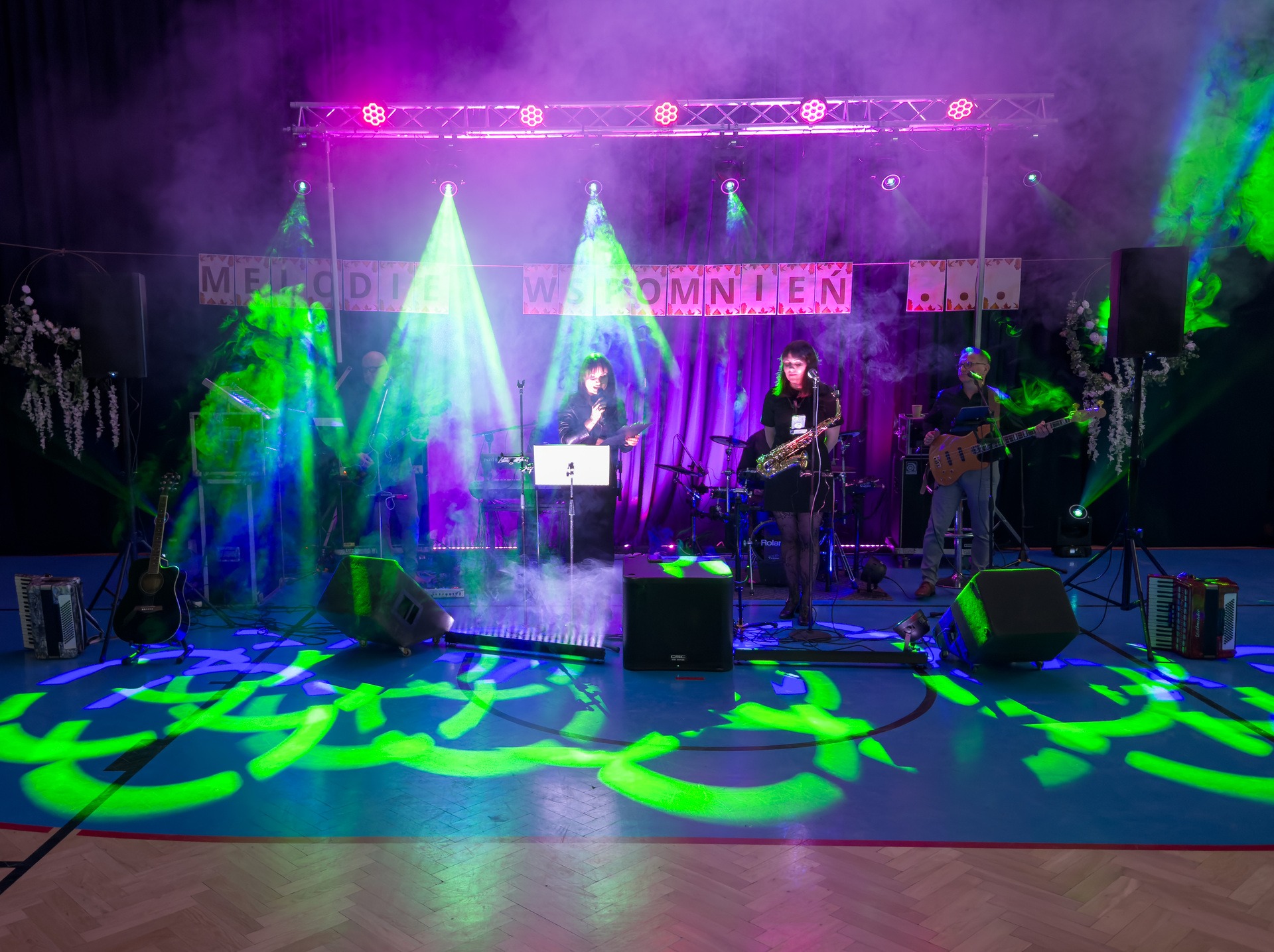 Zespół muzyczny na scenie z oświetleniem i dekoracjami. Wokalistka z saksofonem na pierwszym planie, w tle instrumenty i napis 'Melodie Wspomnień'.