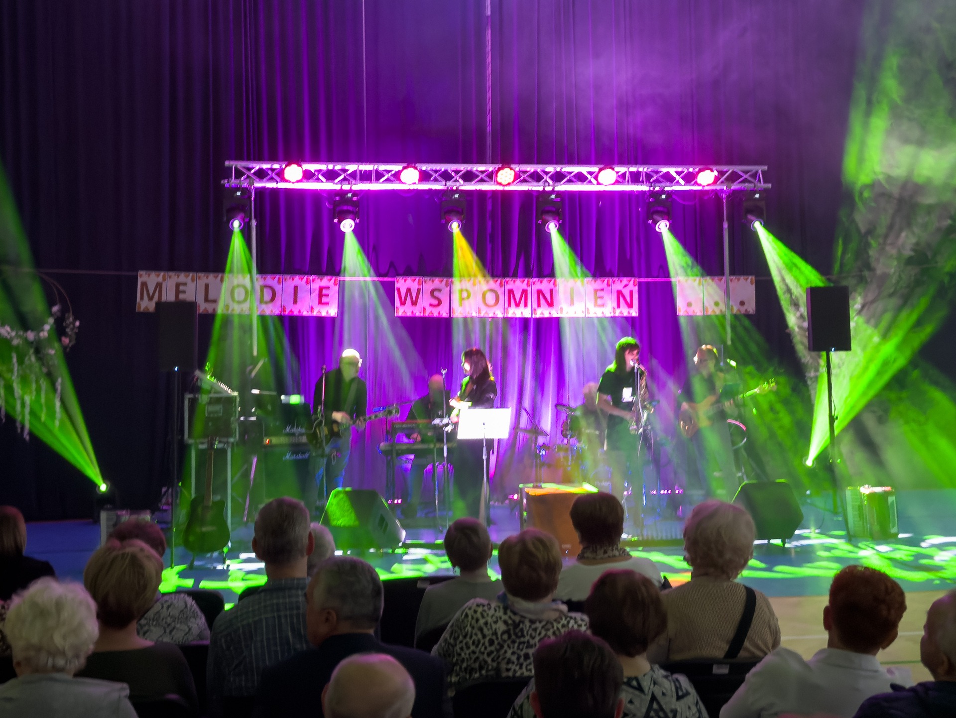 Zespół muzyczny na scenie, oświetlony zielonymi i fioletowymi światłami, gra dla publiczności. Widoczny napis 'Melodie Wspomnień' nad sceną.