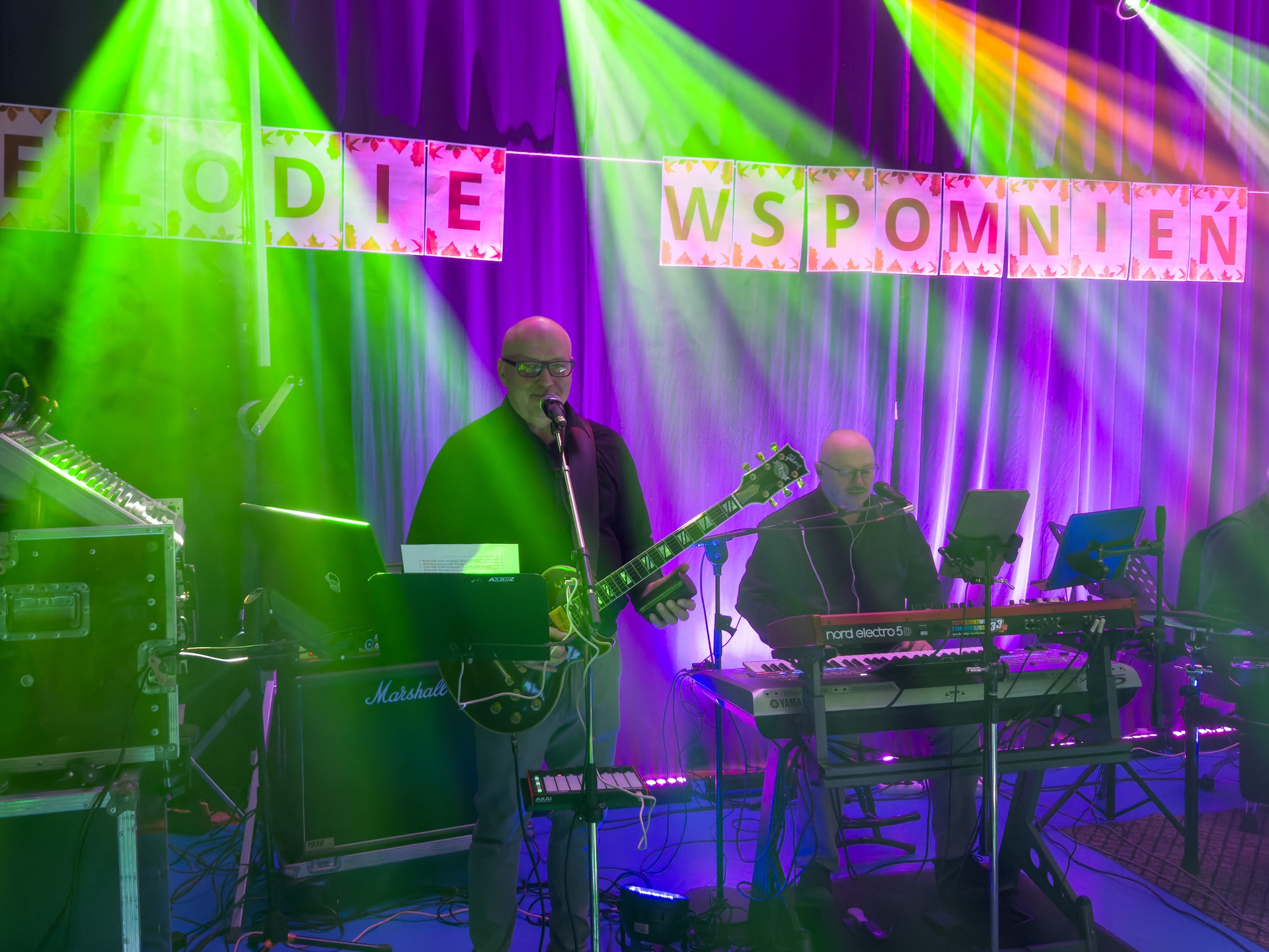 Zespół na scenie z gitarą i keyboardem, podświetlony zielonymi i fioletowymi światłami. Baner z napisem 'Melodie Wspomnień' w tle. Wzmacniacz Marshall.
