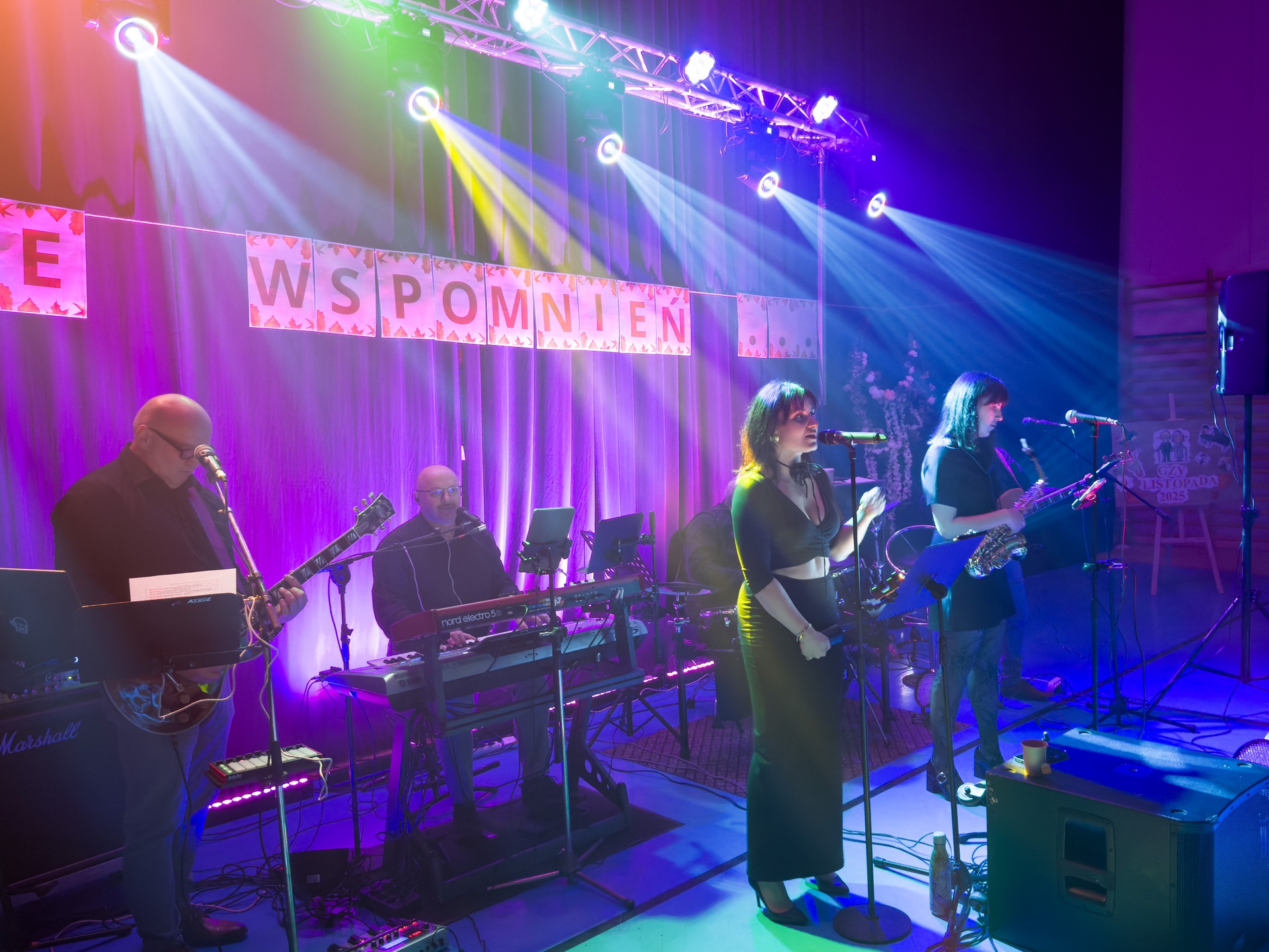 Zespół muzyczny na scenie podczas występu. W tle dekoracja z napisem 'Wspomnienia'. Oświetlenie sceniczne tworzy atmosferę koncertu. Widoczni muzycy grający na instrumentach.