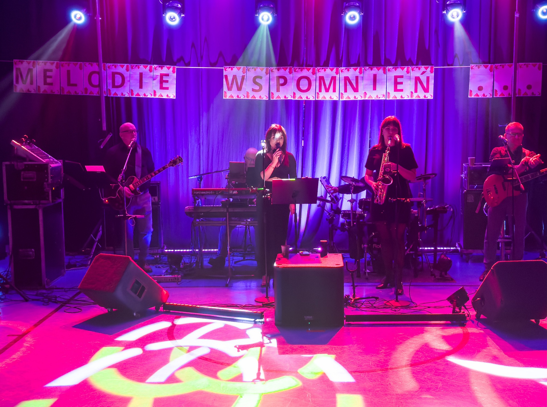 Zespół muzyczny na scenie, oświetlonej fioletowym światłem, z napisem 'Melodie Wspomnień' w tle. Wokalistka śpiewa, a muzycy grają na gitarach, saksofonie i keyboardzie.