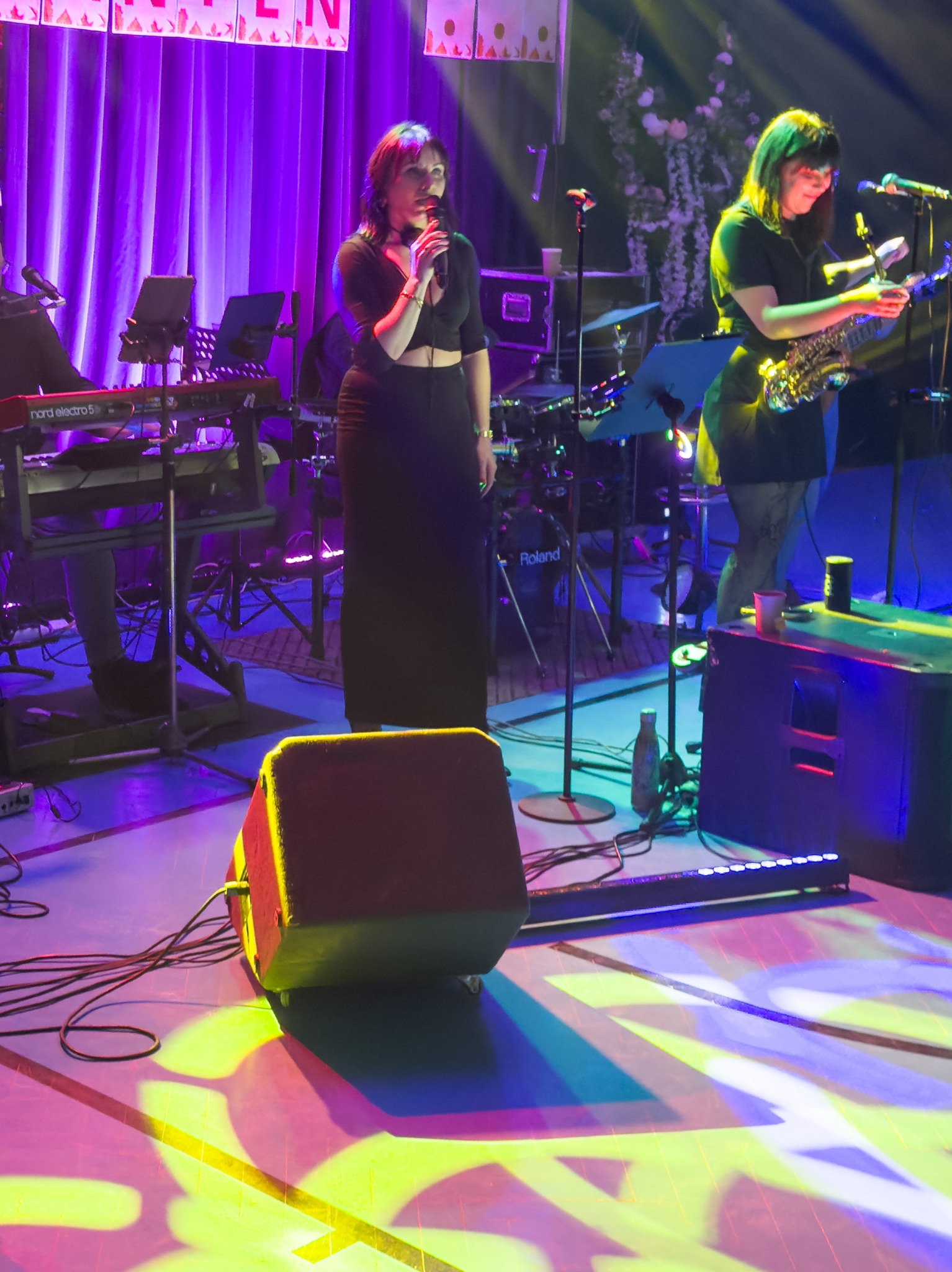 Wokalistka z mikrofonem i saksofonistka na scenie podczas występu. W tle instrumenty, dekoracje kwiatowe i fioletowe światła. Na podłodze kolorowe wzory świetlne.