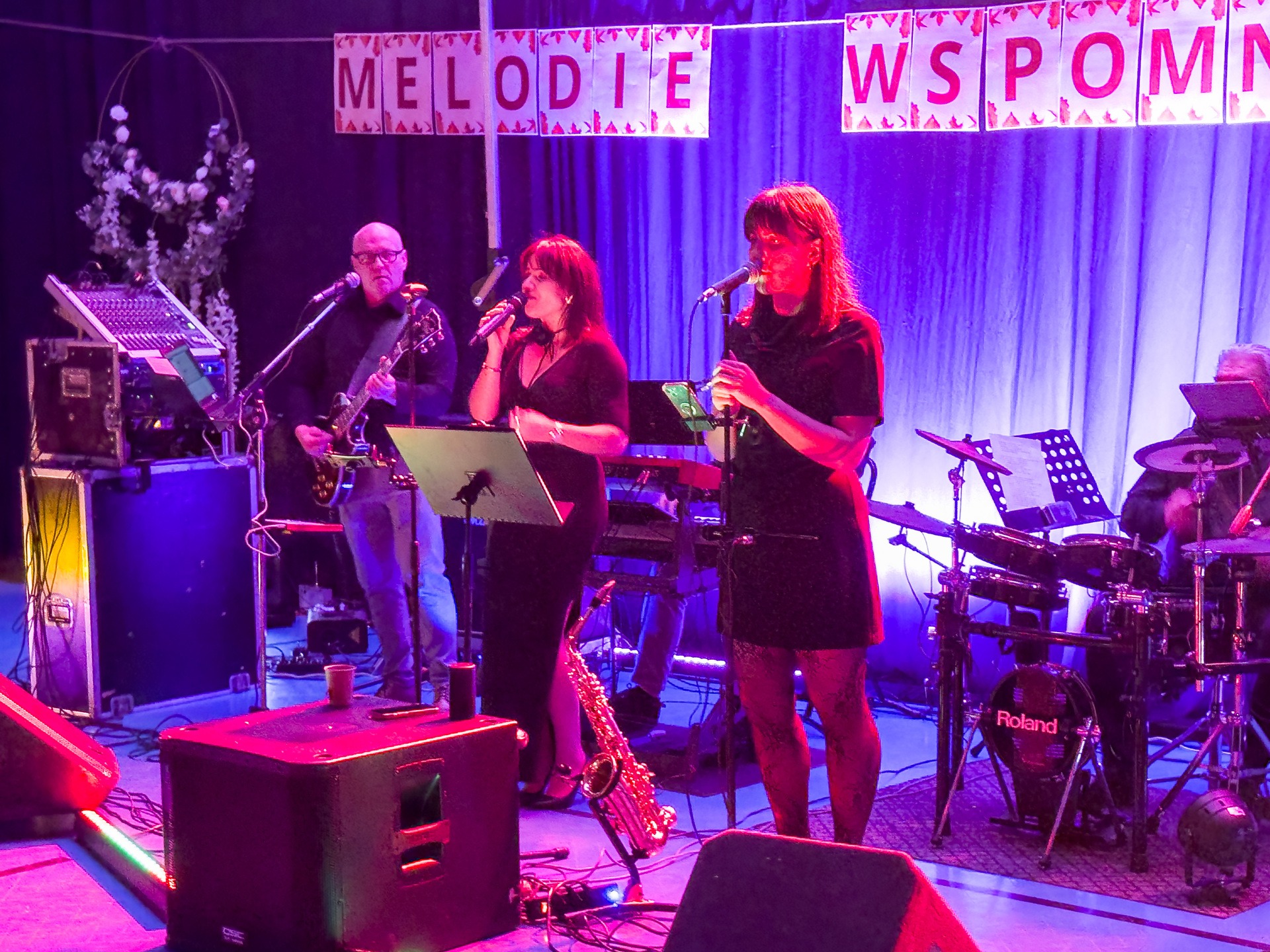 Zespół muzyczny 'Melodie Wspomnień' na scenie: wokalistki z mikrofonami, gitarzysta i perkusista. Scena oświetlona fioletowym światłem, w tle dekoracje.
