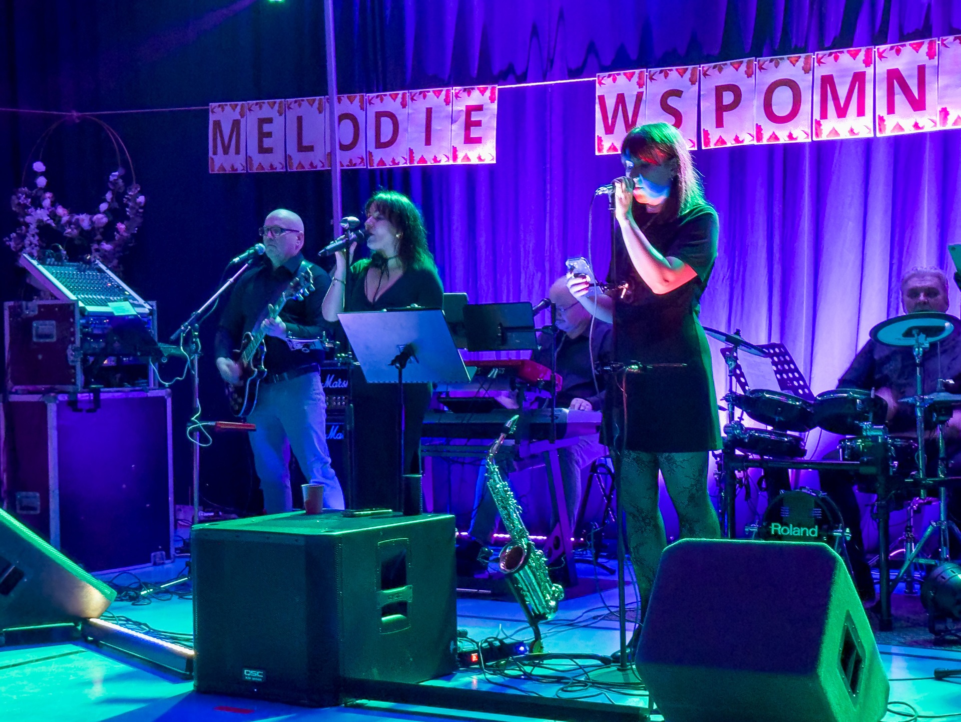 Zespół muzyczny na scenie w trakcie występu, oświetlony niebieskim światłem. Widoczni muzycy grający na instrumentach i wokalistki śpiewające do mikrofonów. Dekoracja z napisem 'Melodie Wspomnień'.