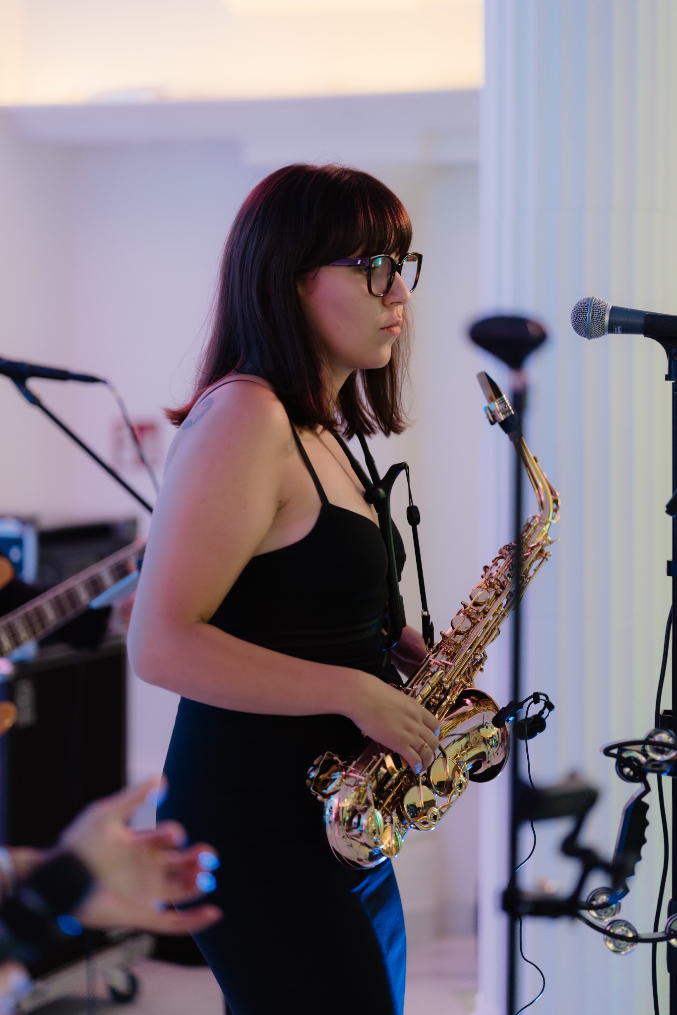 Saksofonistka w czarnej sukience, z okularami, grająca na saksofonie podczas występu na tle kolumny i mikrofonu. W tle widoczna gitara.