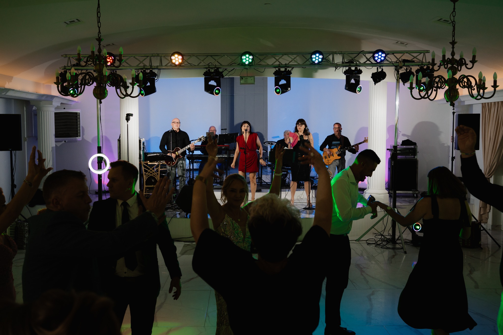 Zespół muzyczny na scenie podczas występu na sali balowej. W tle oświetlenie sceniczne i tańczący goście. Występ uświetniają żyrandole.