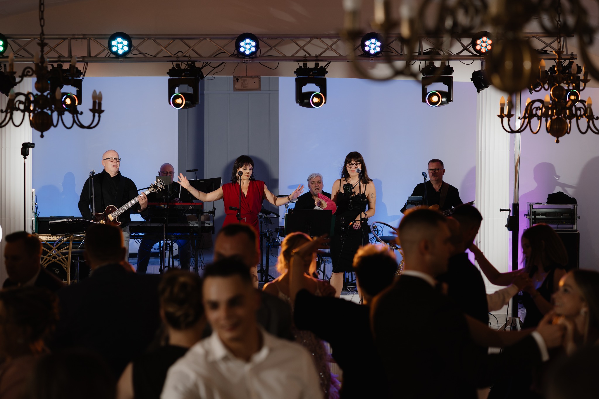 Zespół muzyczny na scenie podczas występu na weselu. W tle oświetlenie sceniczne i tańczący goście. Wokalistki w czerwonej i czarnej sukience, gitarzysta i klawiszowiec.