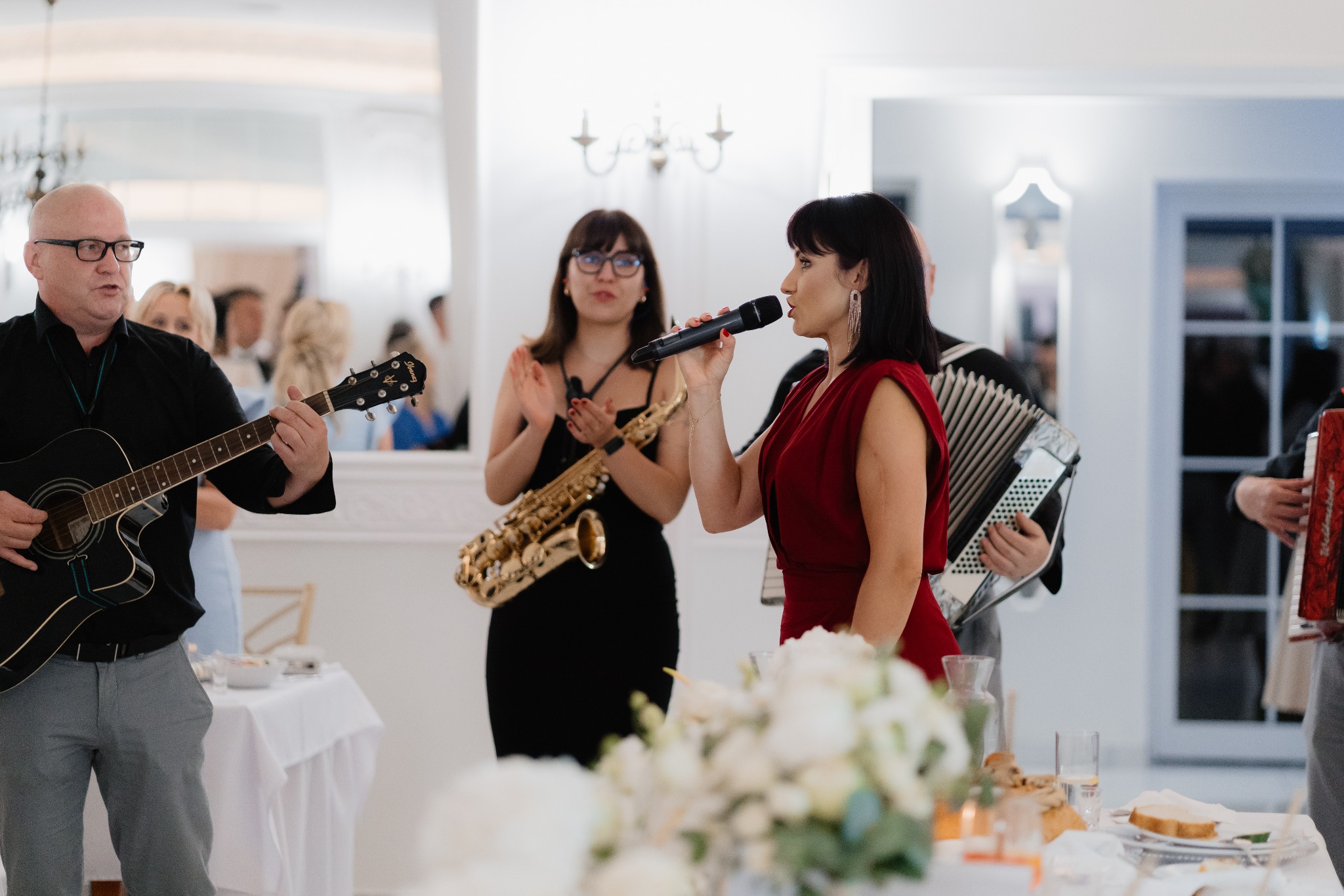 Zespół muzyczny na weselu: wokalistka w czerwonej sukience śpiewa do mikrofonu, obok saksofonistka, gitarzysta i akordeonista. Białe wnętrze sali.
