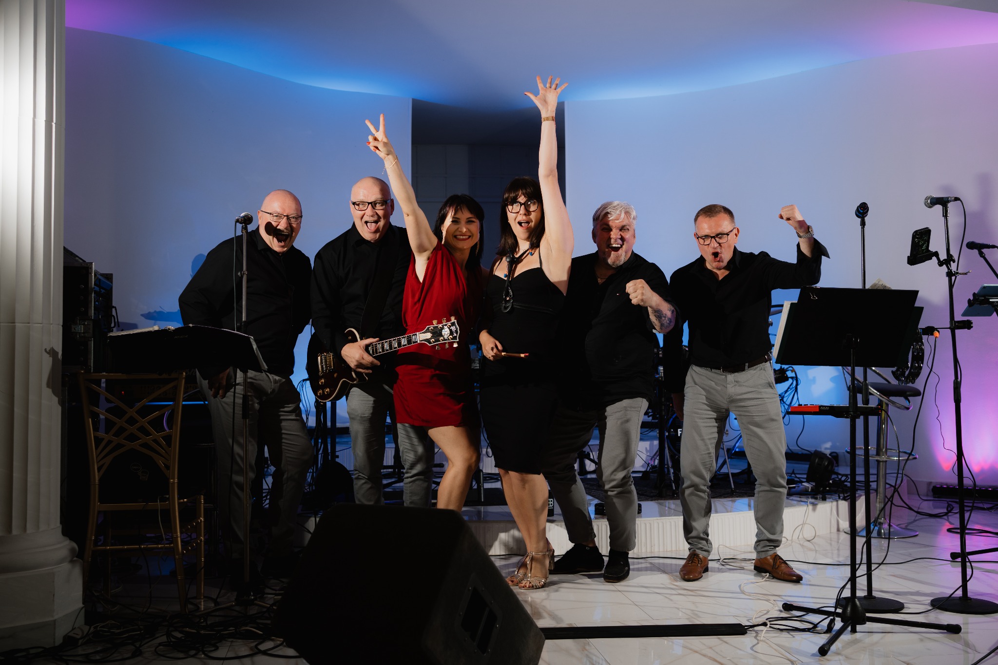 Zespół muzyczny w akcji na scenie z kolorowym oświetleniem. Wokalistki z gitarzystą i perkusistą w radosnych pozach. Widoczne mikrofony, statywy i kolumna głośnikowa.