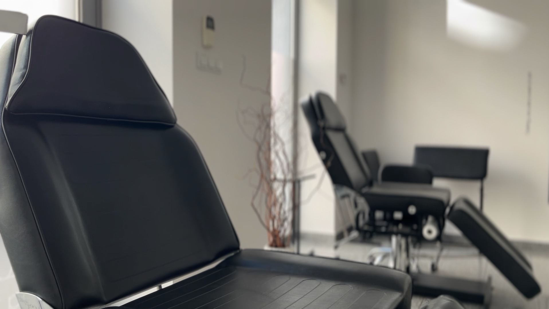 Nowoczesny gabinet kosmetyczny: zbliżenie na czarny fotel zabiegowy z regulacją, w tle drugi fotel i minimalistyczny wystrój w odcieniach szarości.