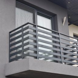 KARBON KAMIL MATANOWSKI - Nowoczesna, szara balustrada balkonowa z poziomych, metalowych profili, zamontowana na balkonie budynku o szarej elewacji. Widoczne okno i fragment dachu.