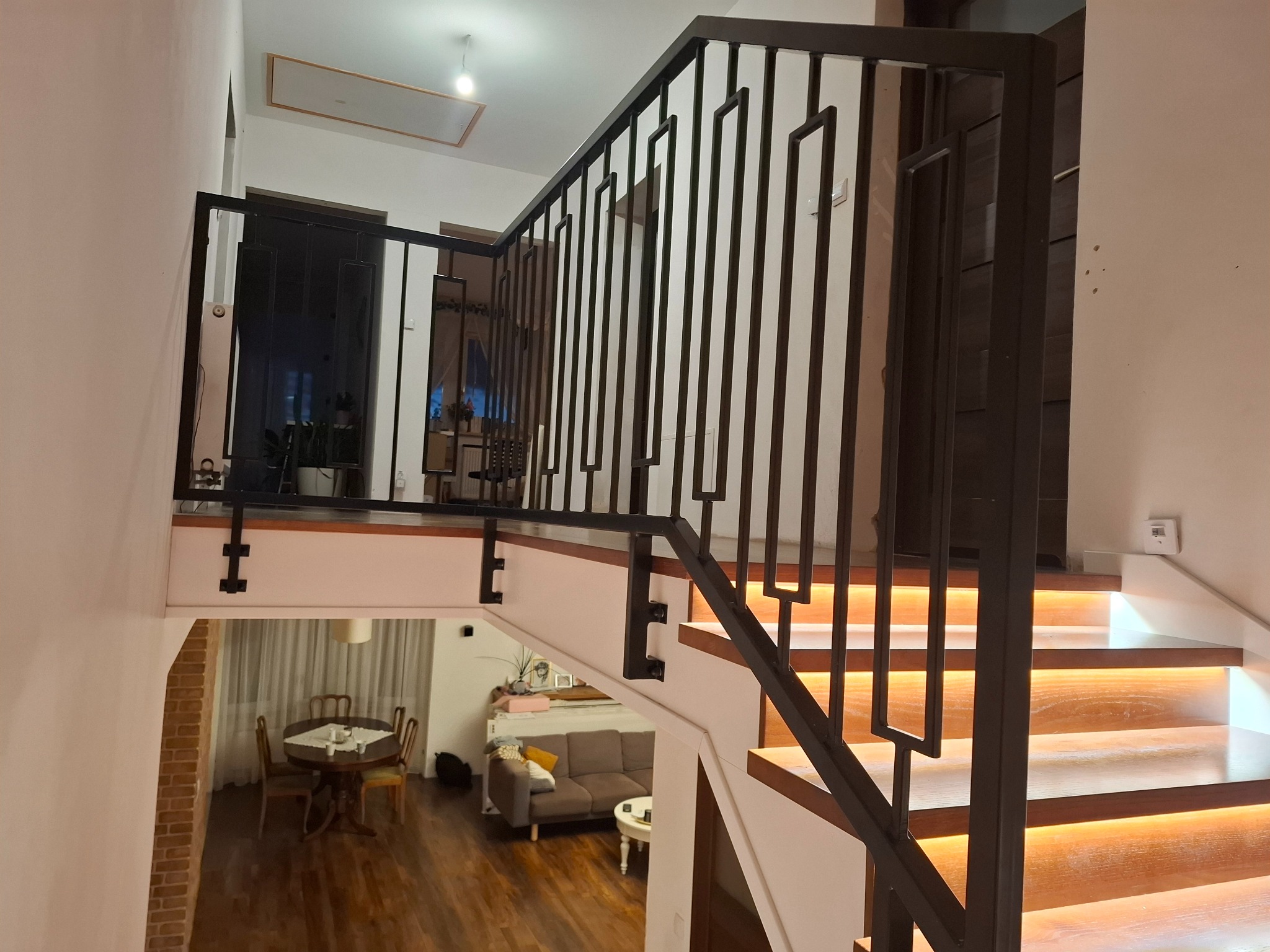 Nowoczesna, czarna balustrada schodowa z geometrycznym wzorem, oświetlone stopnie drewniane. Widok z góry na salon z jadalnią. Minimalistyczny design.
