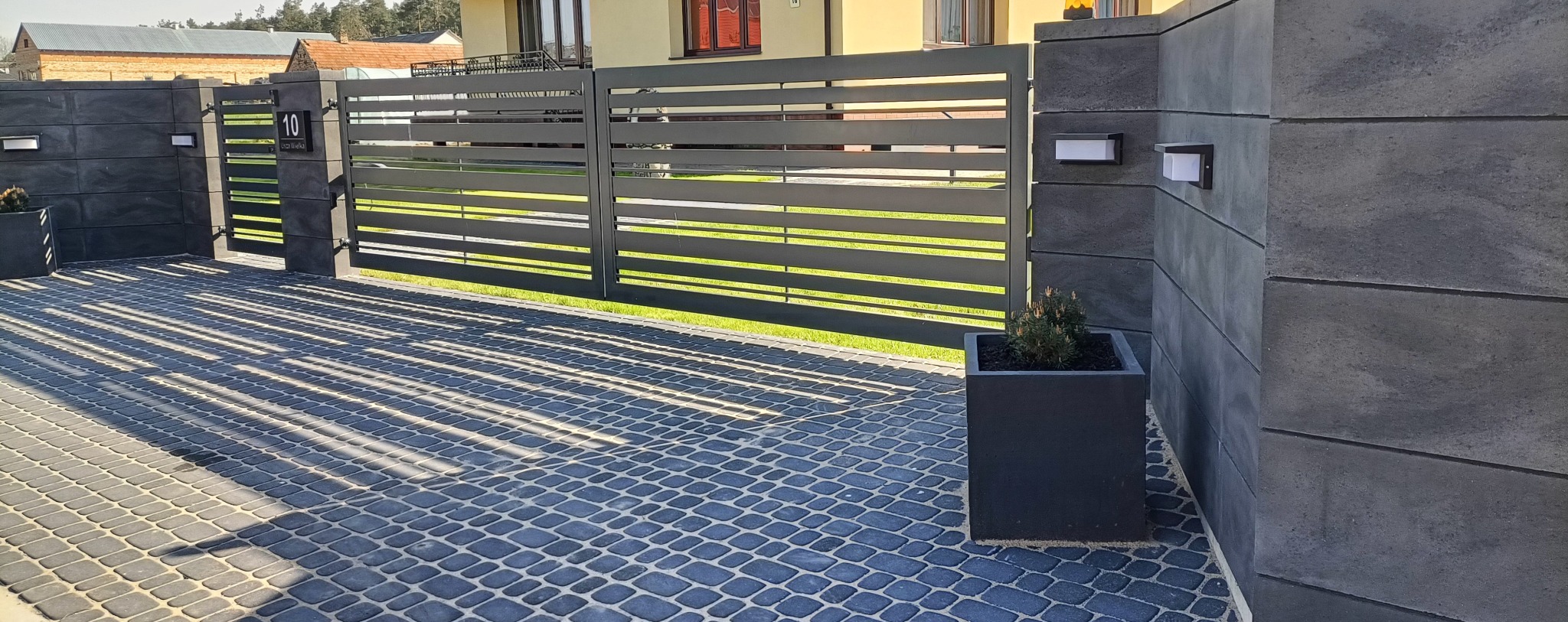 Nowoczesna, pozioma brama przesuwna z szarego metalu, zamontowana na betonowych słupkach z oświetleniem. Kostka brukowa w cieniu bramy. Minimalistyczny design.