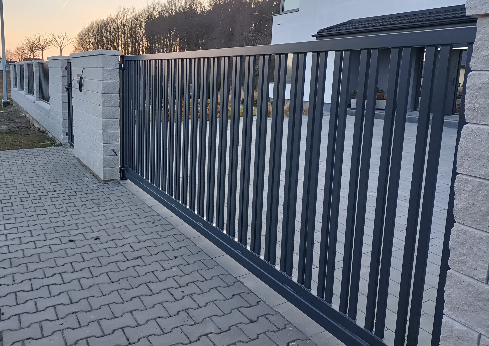 Nowoczesna, przesuwna brama w kolorze antracytu z pionowymi profilami, zamontowana przy posesji z kostką brukową i ogrodzeniem z bloczków betonowych. Minimalistyczny design.