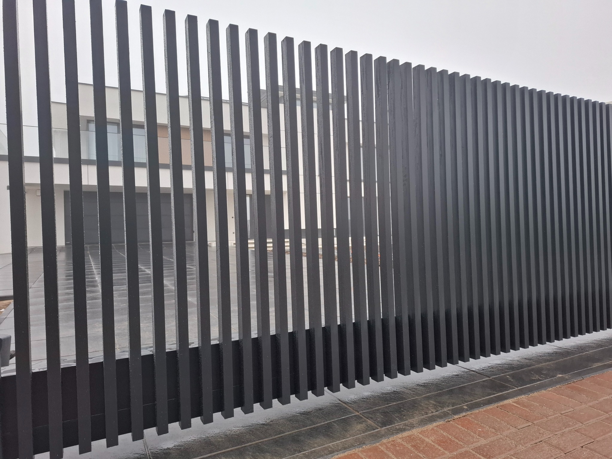 Nowoczesne, grafitowe ogrodzenie panelowe z pionowych profili, na tle jasnego budynku. Minimalistyczny design, widoczne detale wykończenia i fragment chodnika z kostki.