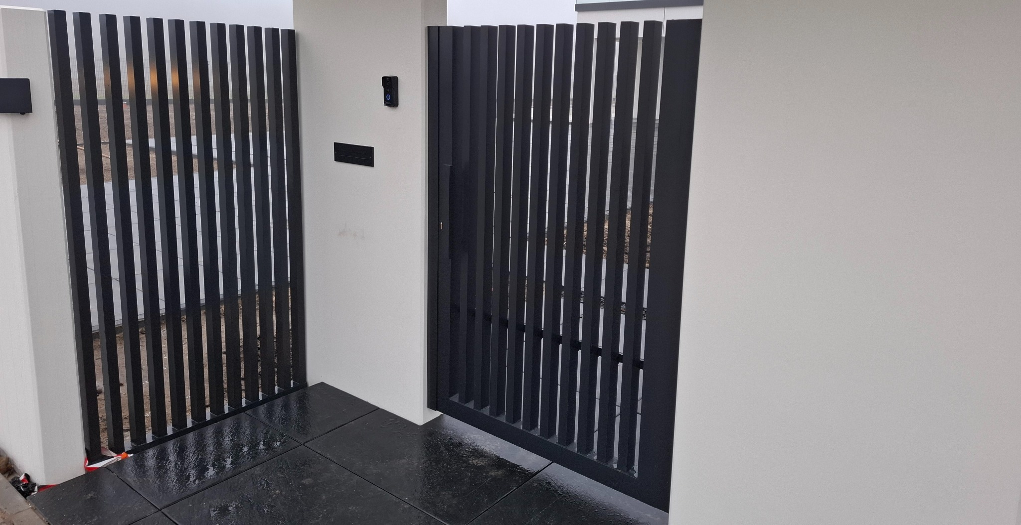 Nowoczesna, czarna brama metalowa z pionowymi szczebelkami, wkomponowana w białe, minimalistyczne ogrodzenie. Widoczny fragment czarnej kostki brukowej.