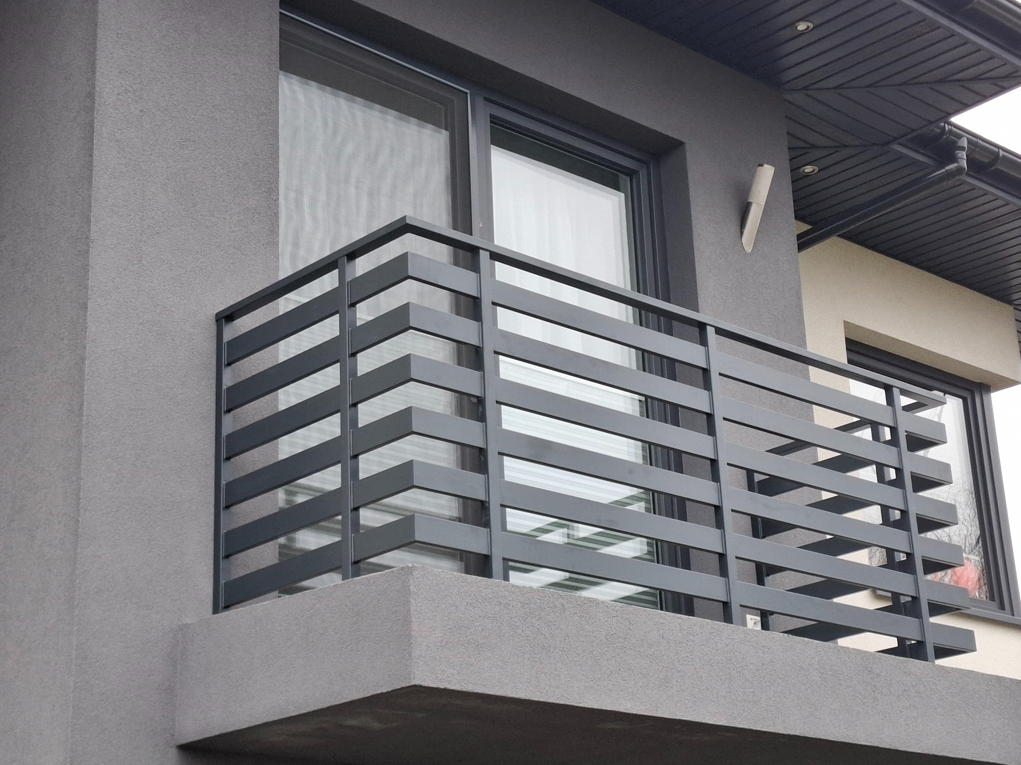 Nowoczesna, szara balustrada balkonowa z poziomych, metalowych profili, zamontowana na balkonie budynku o szarej elewacji. Widoczne okno i fragment dachu.