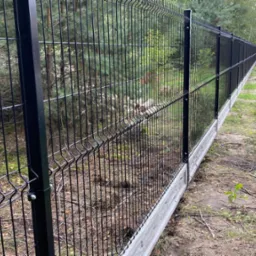 Długi, czarny panelowy płot z podmurówką z betonu, zamontowany wzdłuż granicy działki, z widocznymi drzewami w tle. Perspektywa z boku, ukazuje detale konstrukcyjne.