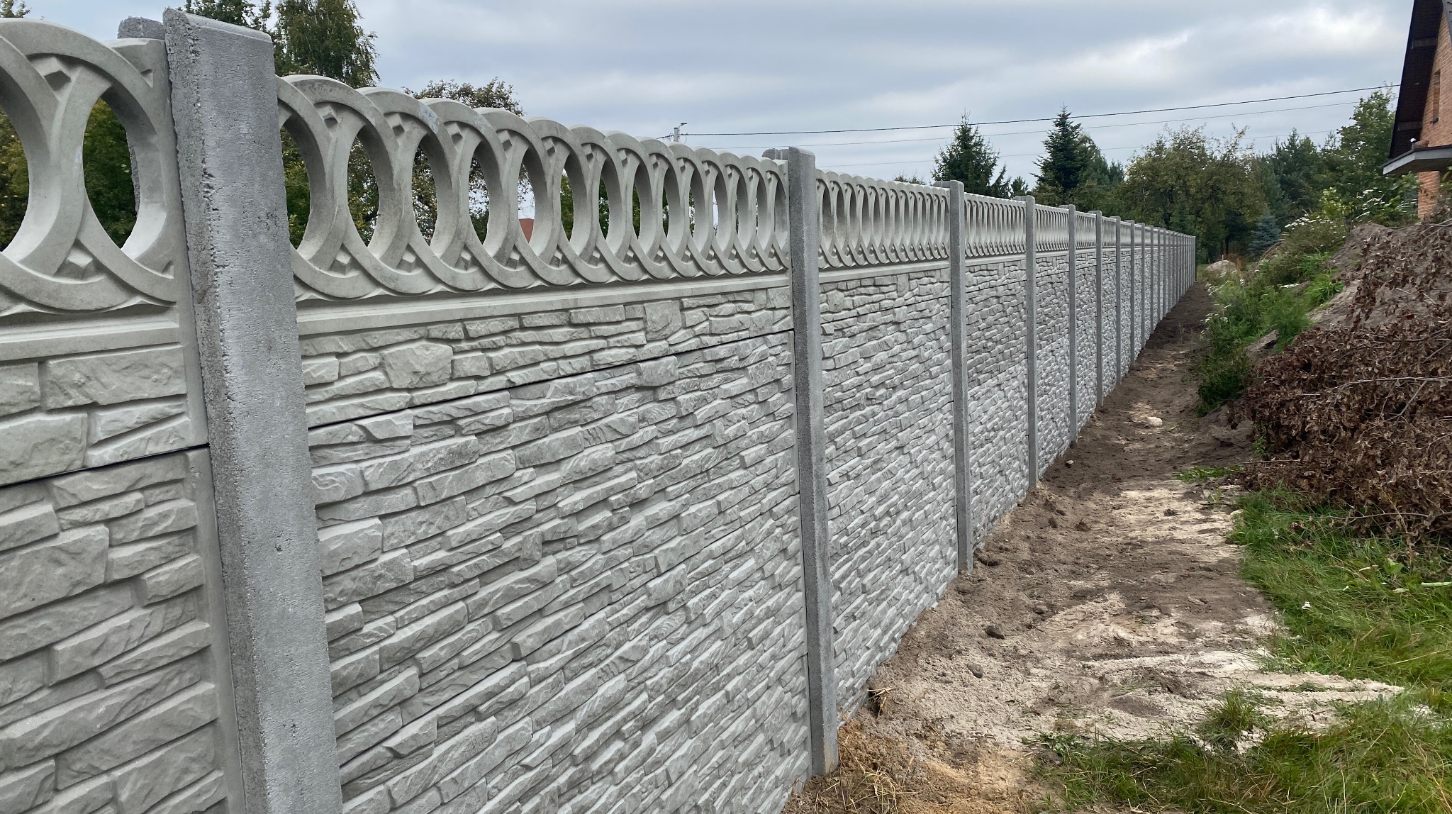 Długi, szary, betonowy płot z dekoracyjnymi okręgami u góry, imitujący kamień, oddziela posesję. Widok z perspektywy ziemi, na tle zieleni i pochmurnego nieba.
