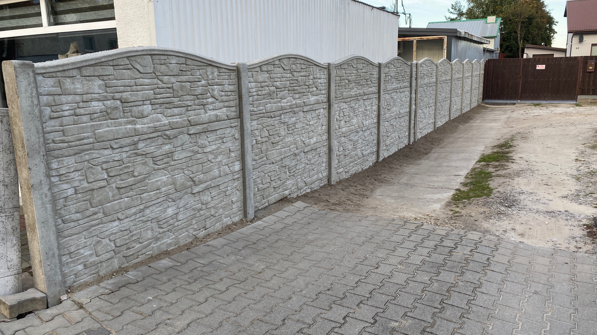 Długi, szary, betonowy płot z dekoracyjnym wzorem kamienia, oddzielający posesję od drogi z kostki brukowej. Widok z perspektywy.
