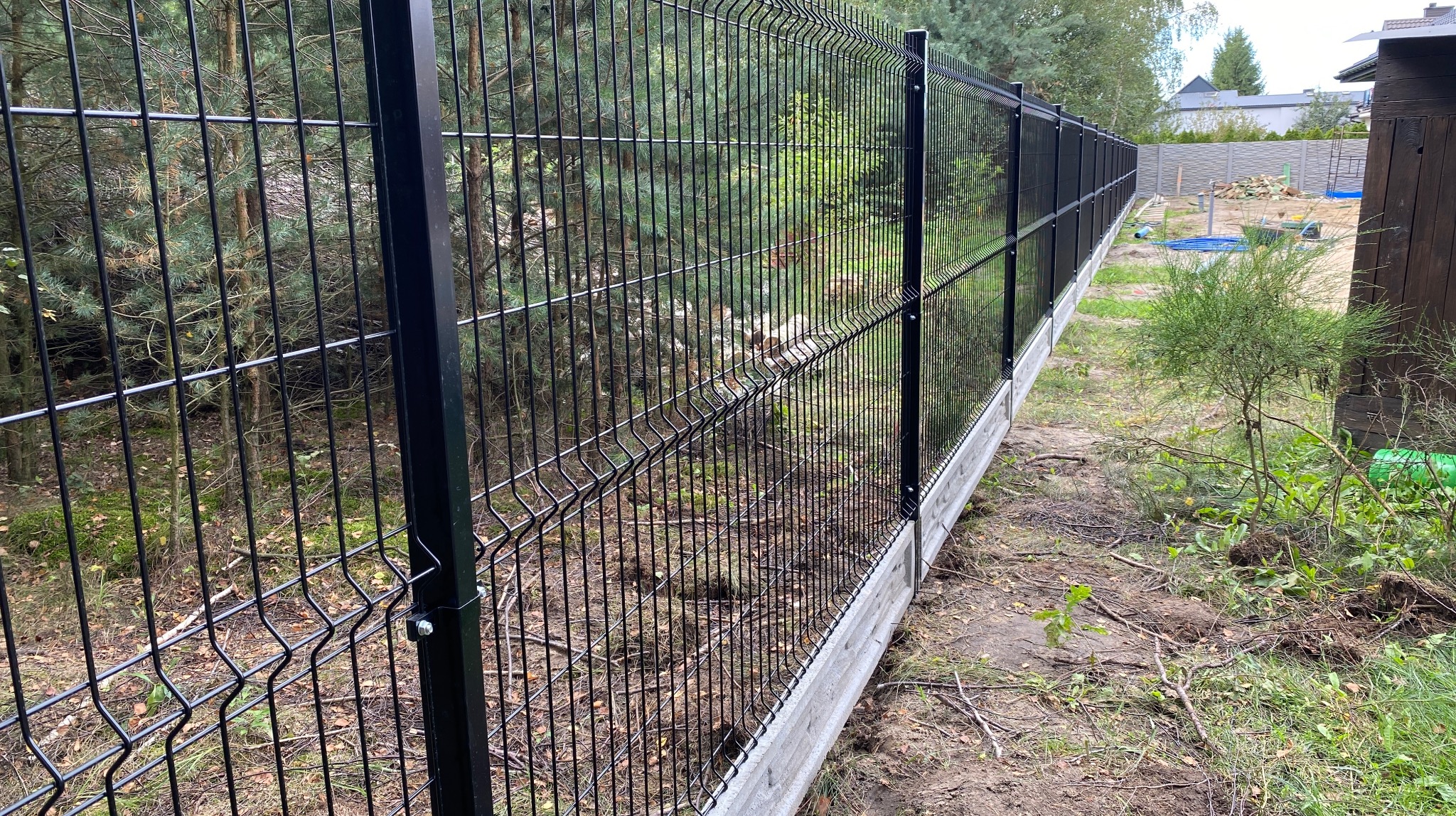 Długi, czarny panelowy płot z podmurówką z betonu, zamontowany wzdłuż granicy działki, z widocznymi drzewami w tle. Perspektywa z boku, ukazuje detale konstrukcyjne.