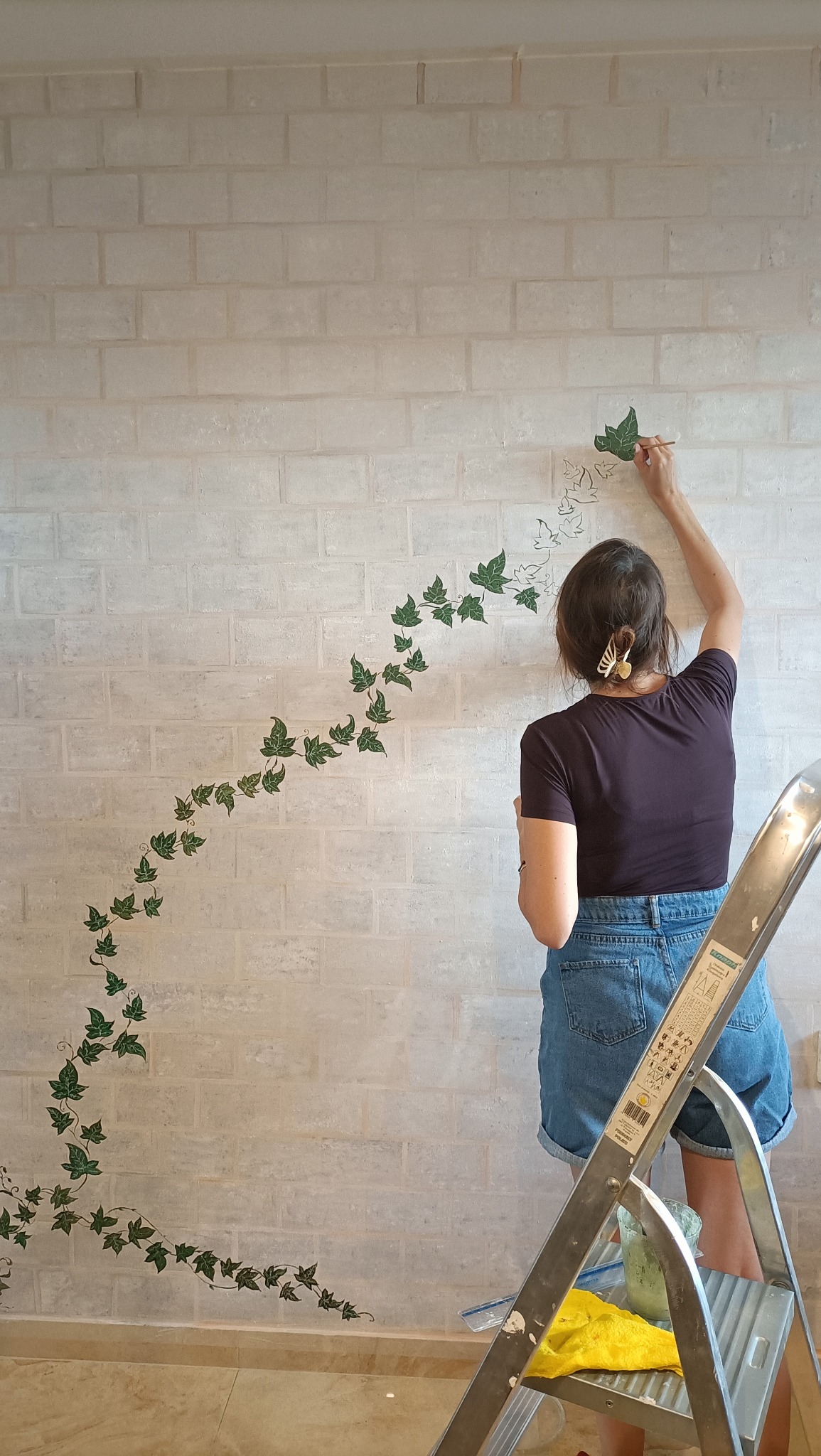 Artystyczne malowanie ściany z cegieł: kobieta z pędzlem dodaje zielone motywy roślinne, tworząc dekoracyjny wzór pnącza. Drabina i farba w tle.