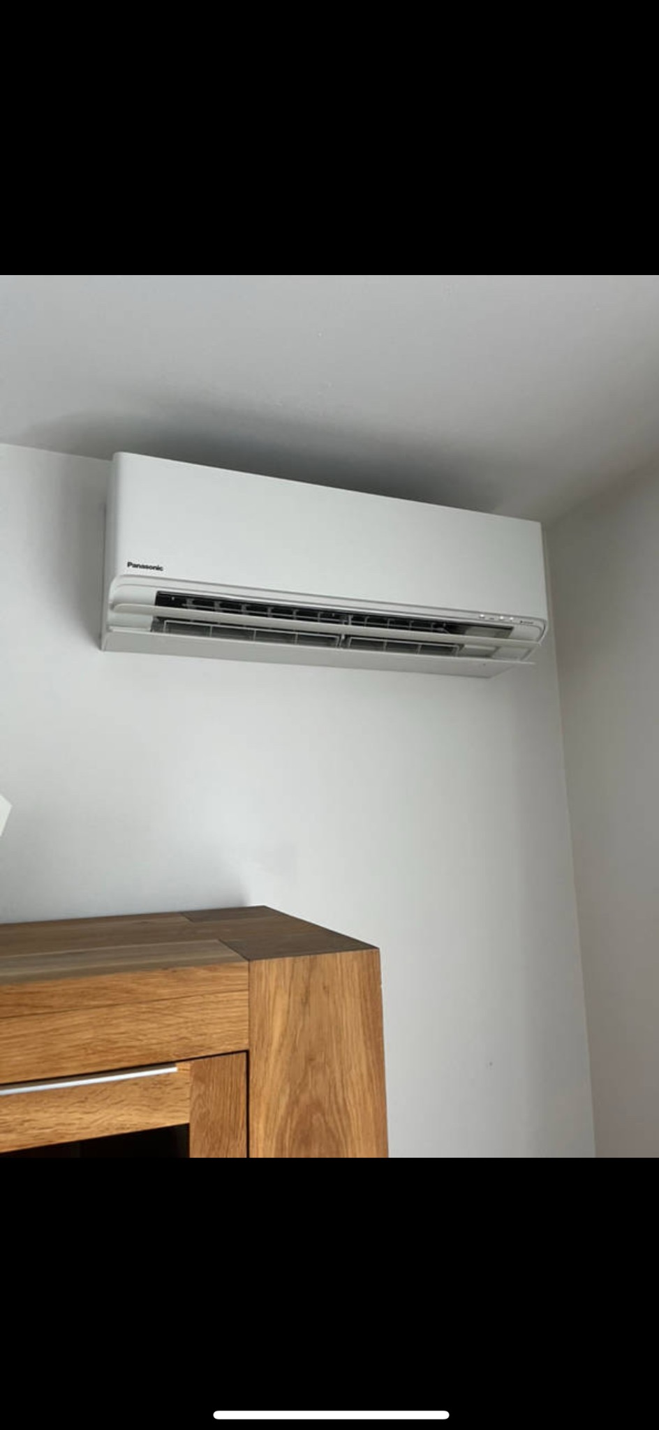 Biały klimatyzator Panasonic zamontowany na ścianie nad drewnianą komodą. Widok z dołu, oświetlenie dzienne.