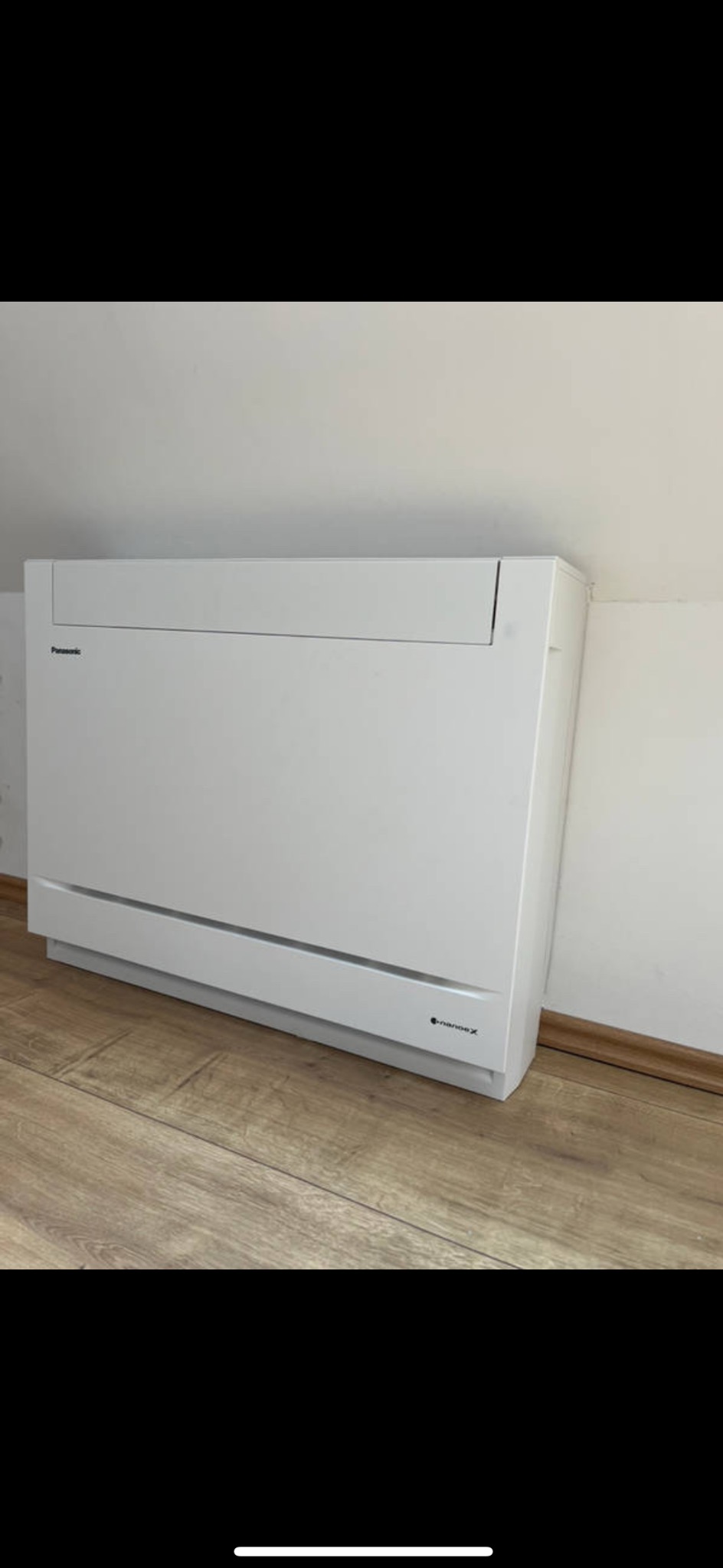 Biały klimatyzator podłogowy Panasonic Nanoe X na drewnianej podłodze, przy białej ścianie. Minimalistyczny design, nowoczesne rozwiązanie grzewcze.