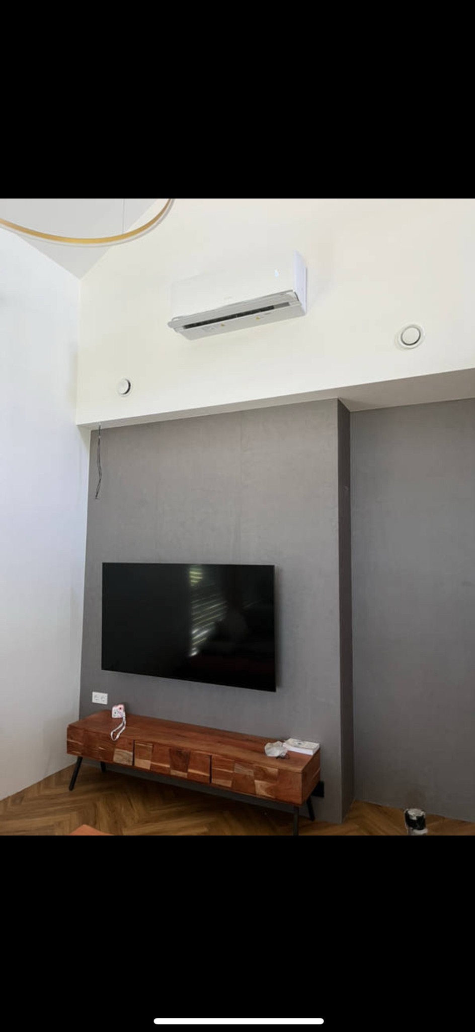 Nowoczesny salon z klimatyzacją ścienną, szarą ścianą i drewnianą szafką RTV. Widoczne elementy instalacji wentylacyjnej i oświetlenie sufitowe.