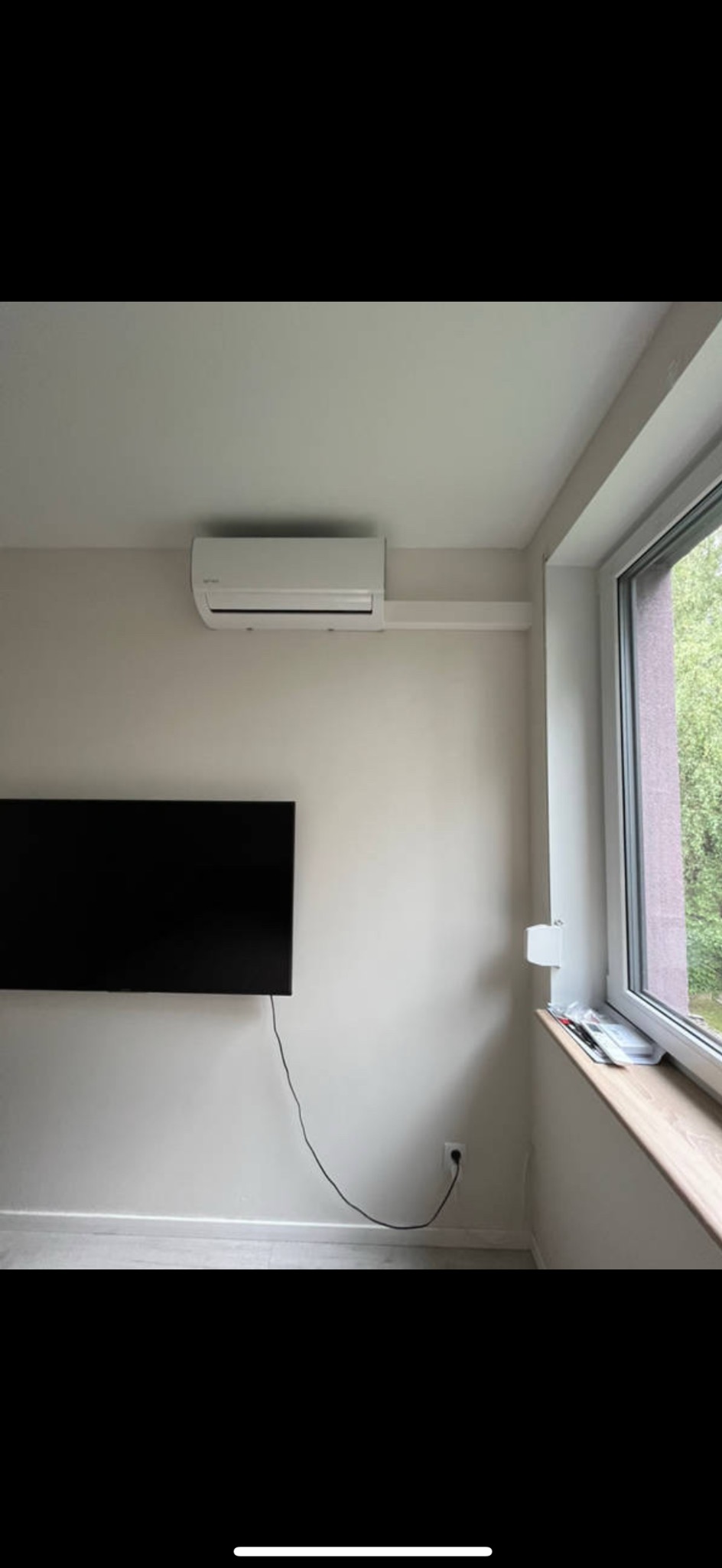 Biały klimatyzator ścienny zamontowany w jasnym pokoju z telewizorem i oknem. Widoczne przewody i gniazdko elektryczne. Minimalistyczny design.