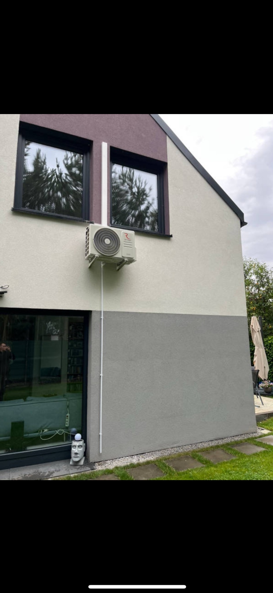 Biała jednostka zewnętrzna klimatyzacji zamontowana na elewacji dwukondygnacyjnego domu z szarą dolną i beżową górną częścią. Widoczne okna z czarnymi ramami i biała rura.