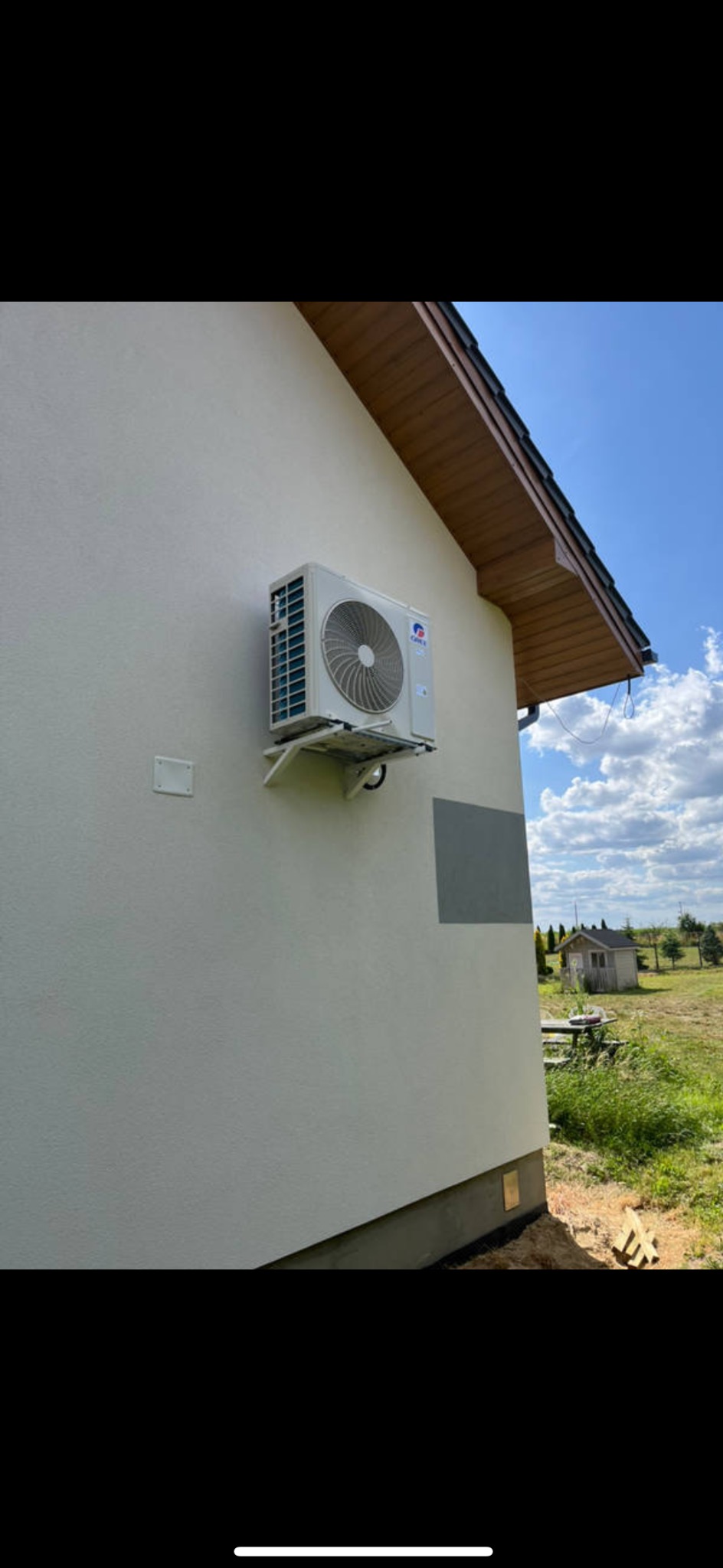 Biały klimatyzator marki Gree zamontowany na elewacji domu z widocznym wspornikiem, na tle błękitnego nieba i zieleni. Widoczny fragment dachu z brązową podbitką.