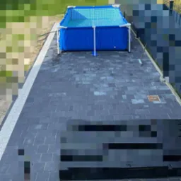 Ułożona szara kostka brukowa z białym obramowaniem, na której stoi niebieski basen. Widok z góry, zieleń w tle.