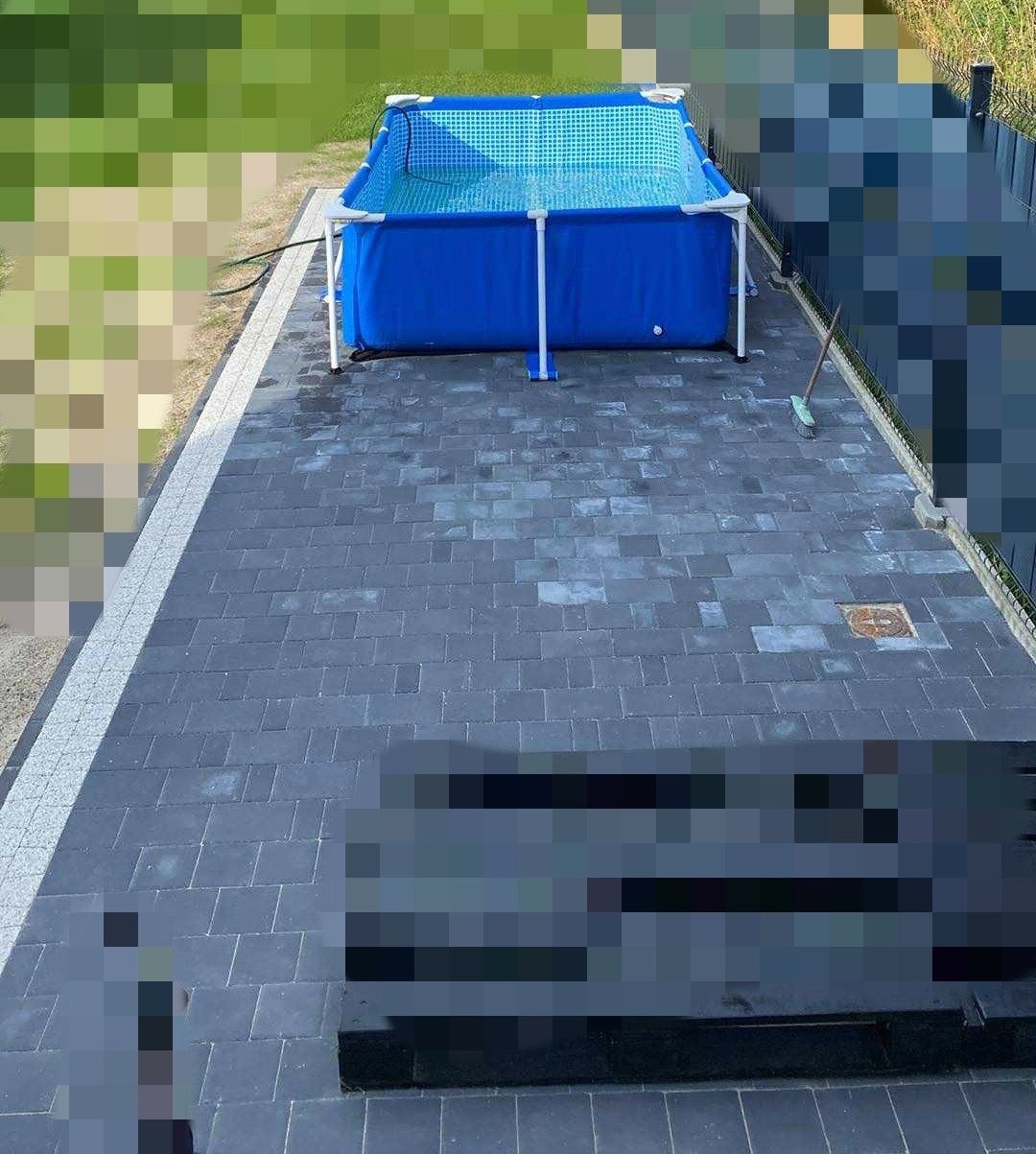 Ułożona szara kostka brukowa z białym obramowaniem, na której stoi niebieski basen. Widok z góry, zieleń w tle.