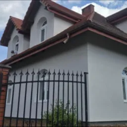 Wykończona elewacja domu z czerwonym dachem i oknami w kształcie łuku. Widoczny fragment ogrodzenia z kutego żelaza i ceglane filary.