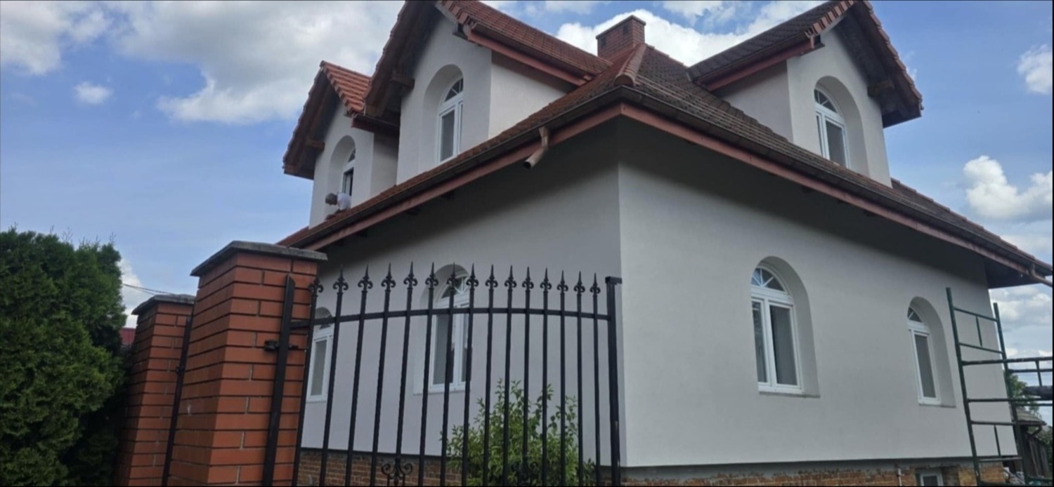 Wykończona elewacja domu z czerwonym dachem i oknami w kształcie łuku. Widoczny fragment ogrodzenia z kutego żelaza i ceglane filary.