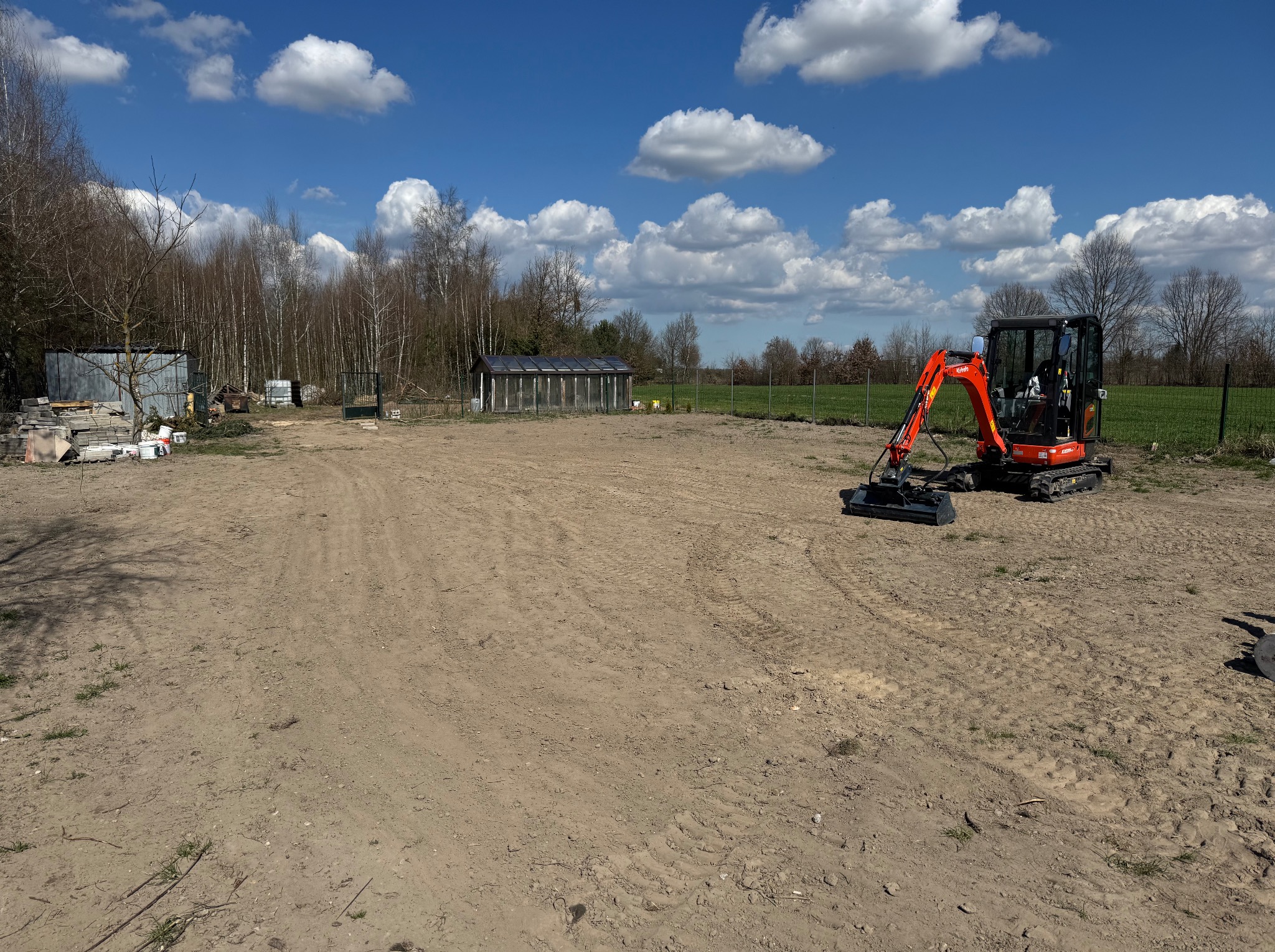 Pomarańczowa minikoparka Kubota na wyrównanym terenie pod budowę, w tle zielony trawnik, drzewa i błękitne niebo z chmurami. Widoczne ślady opon na piasku.