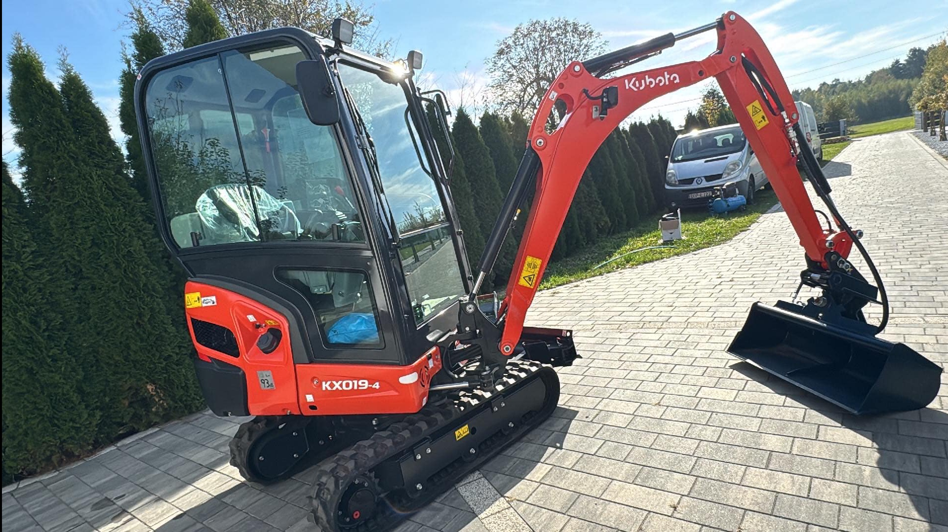 Czerwona mini koparka Kubota KX019-4 na brukowanym podłożu, z widoczną kabiną i łyżką. W tle zaparkowany bus i zieleń drzew.