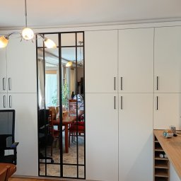 Usługi Stolarskie - Biała szafa wnękowa z lustrem w czarnej ramie, widok na jadalnię. Minimalistyczny design, jasne wnętrze, drewniana podłoga i dywan w odcieniach szarości.