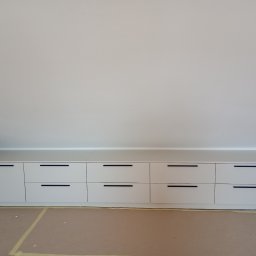 Usługi Stolarskie - Biała zabudowa meblowa ze szufladami pod skosem dachu, z minimalistycznymi czarnymi uchwytami, na podłodze zabezpieczonej kartonem.