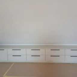 Biała zabudowa meblowa ze szufladami pod skosem dachu, z minimalistycznymi czarnymi uchwytami, na podłodze zabezpieczonej kartonem.