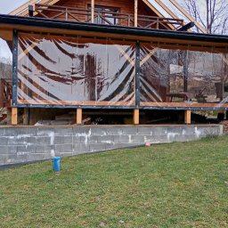 Usługi Stolarskie - Zadaszony taras drewniany z przezroczystymi roletami, widok z zewnątrz na tle trawnika. Konstrukcja z bali, zadaszenie z widoczną konstrukcją więźby dachowej.