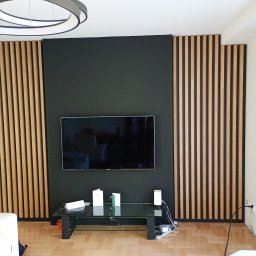 Usługi Stolarskie - Nowoczesny salon z drewnianymi lamelami na ścianie, czarnym tłem pod telewizorem i minimalistycznym oświetleniem. Widoczny fragment podłogi z parkietu.