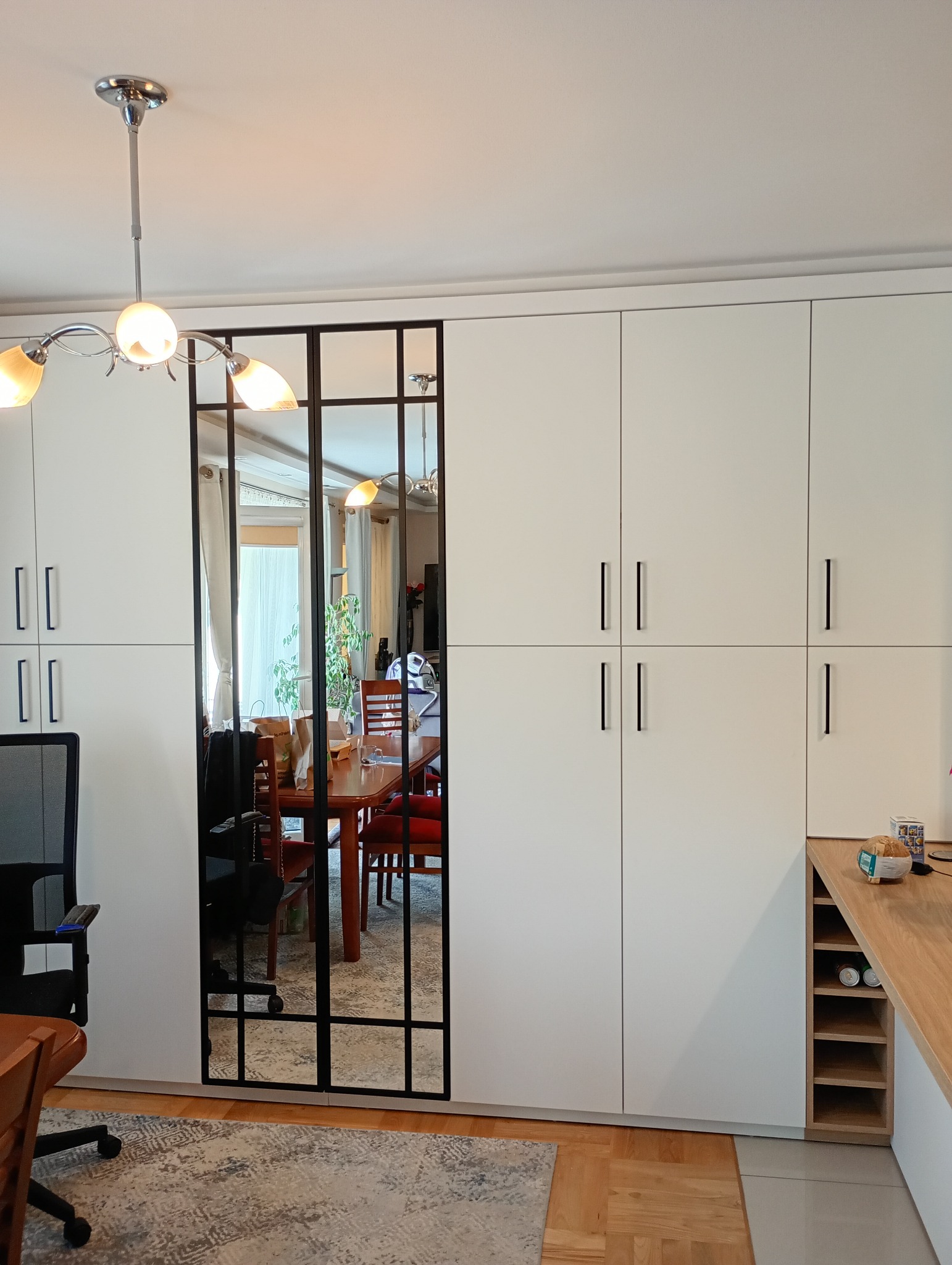 Biała szafa wnękowa z lustrem w czarnej ramie, widok na jadalnię. Minimalistyczny design, jasne wnętrze, drewniana podłoga i dywan w odcieniach szarości.